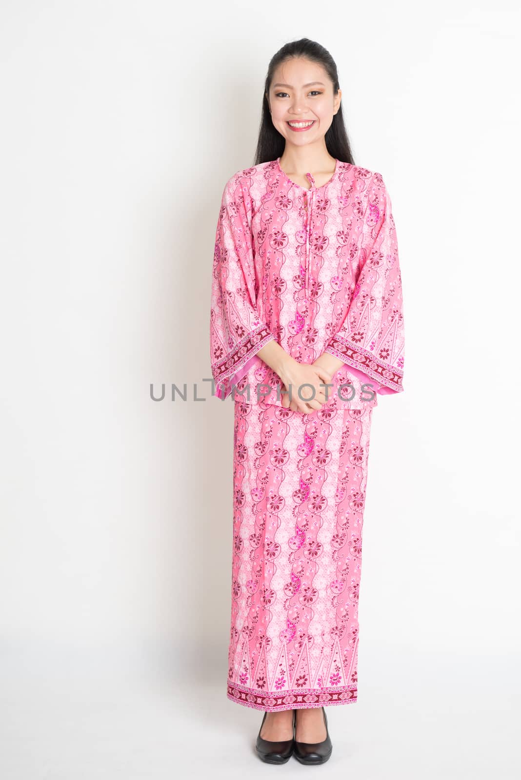 Full body portrait of happy Southeast Asian girl in pink batik dress standing on plain background.