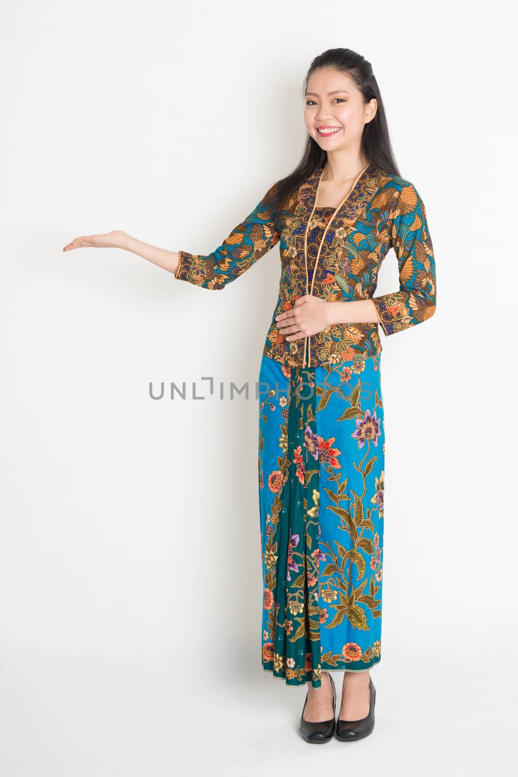 Full length portrait of Southeast Asian woman in batik dress hand holding something standing on plain background.