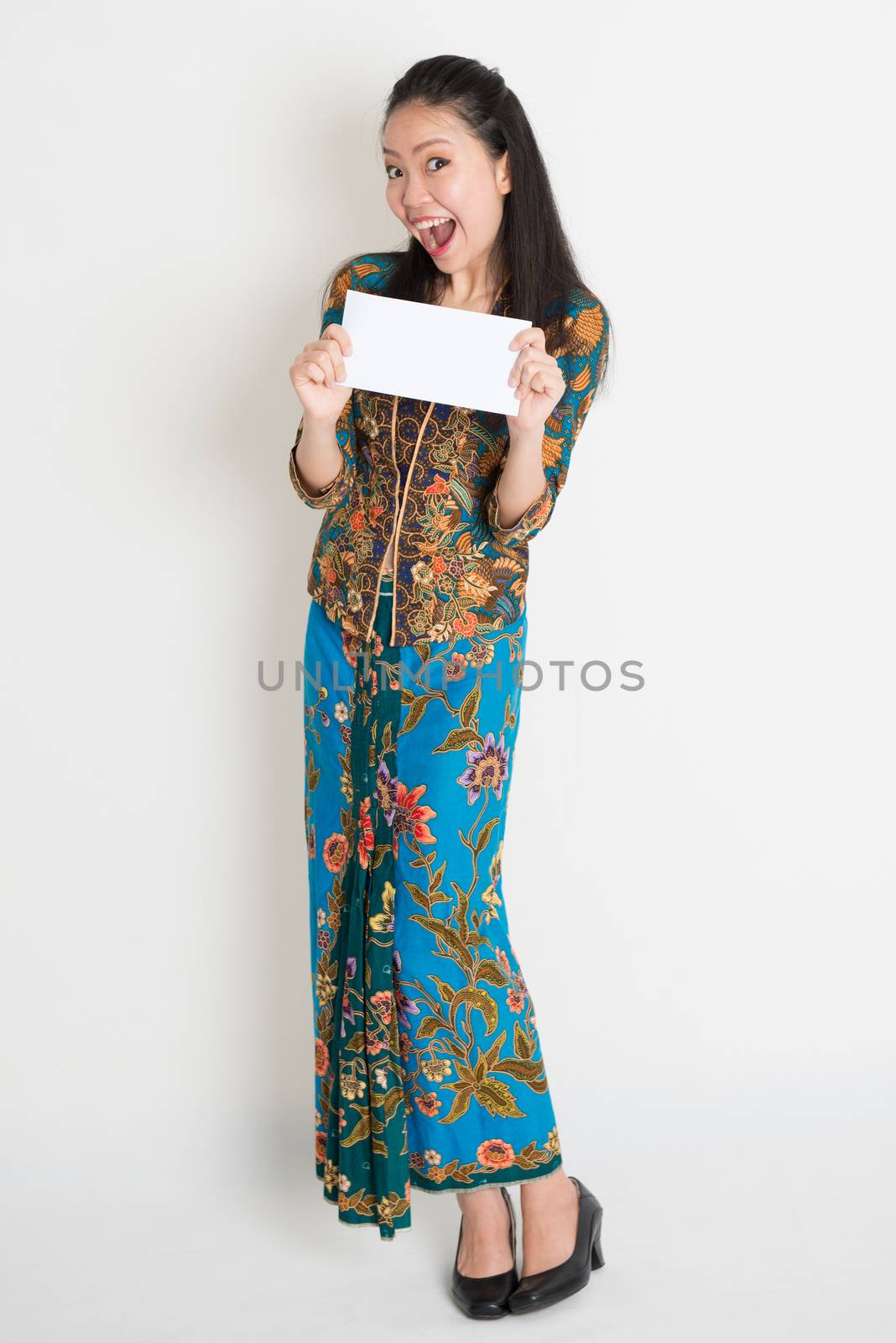 Full length portrait of excited Southeast Asian female in batik dress hands holding an envelope standing on plain background.