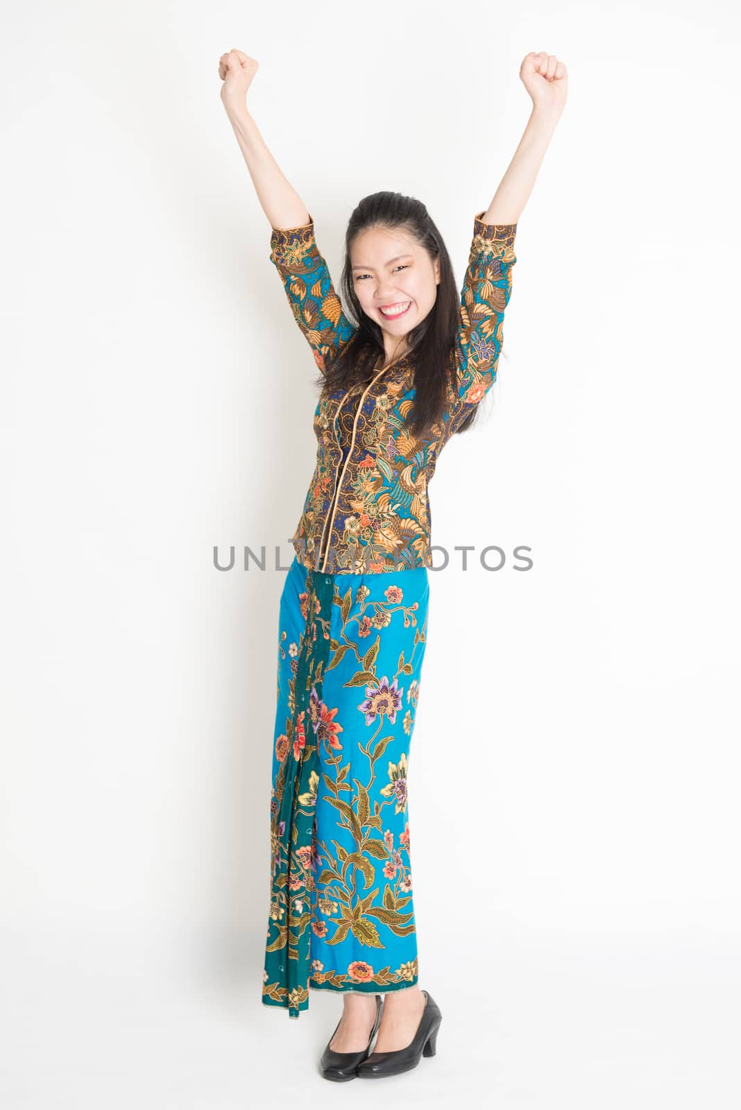 Full length portrait of Southeast Asian female in batik dress arms up and cheering, standing on plain background.