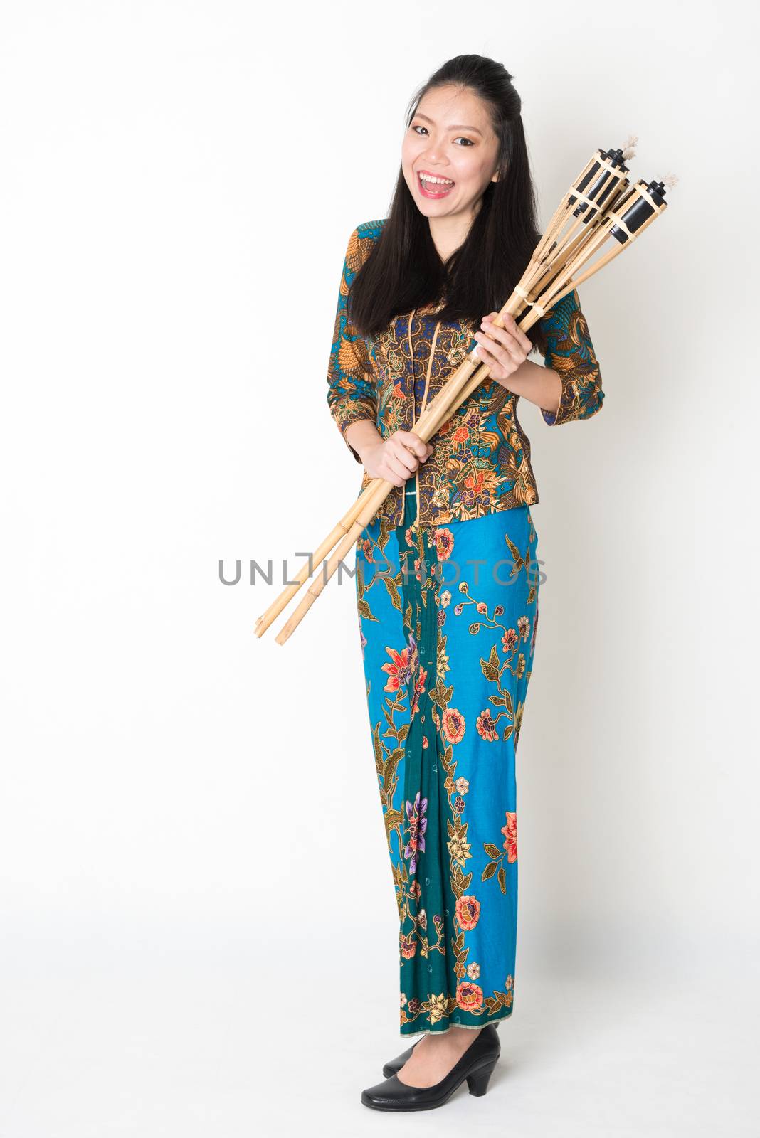 Full body portrait of Southeast Asian girl in batik dress hands holding bamboo torch standing on plain background.