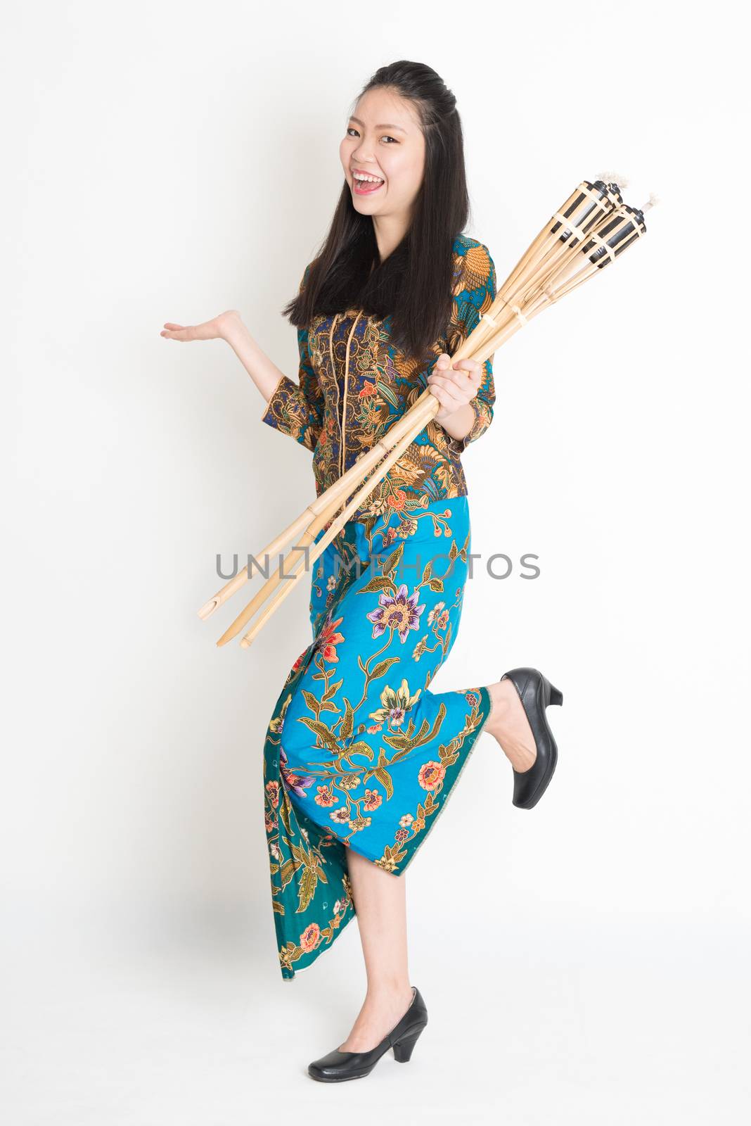 Full body portrait of happy Southeast Asian woman in batik dress hand holding bamboo oil lamp standing on plain background.