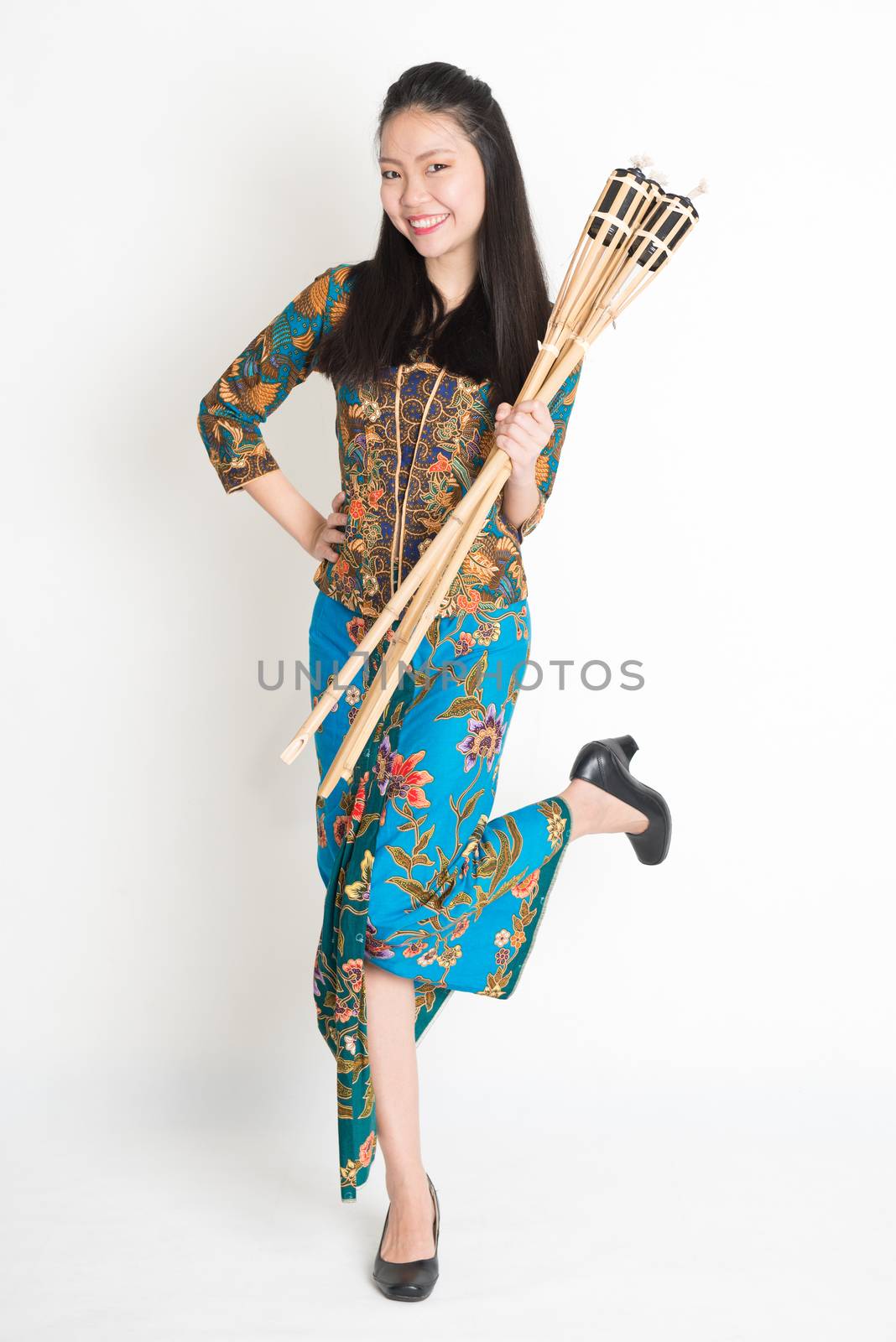 Full body portrait of Southeast Asian woman in batik dress hands holding bamboo oil lamp standing on plain background.