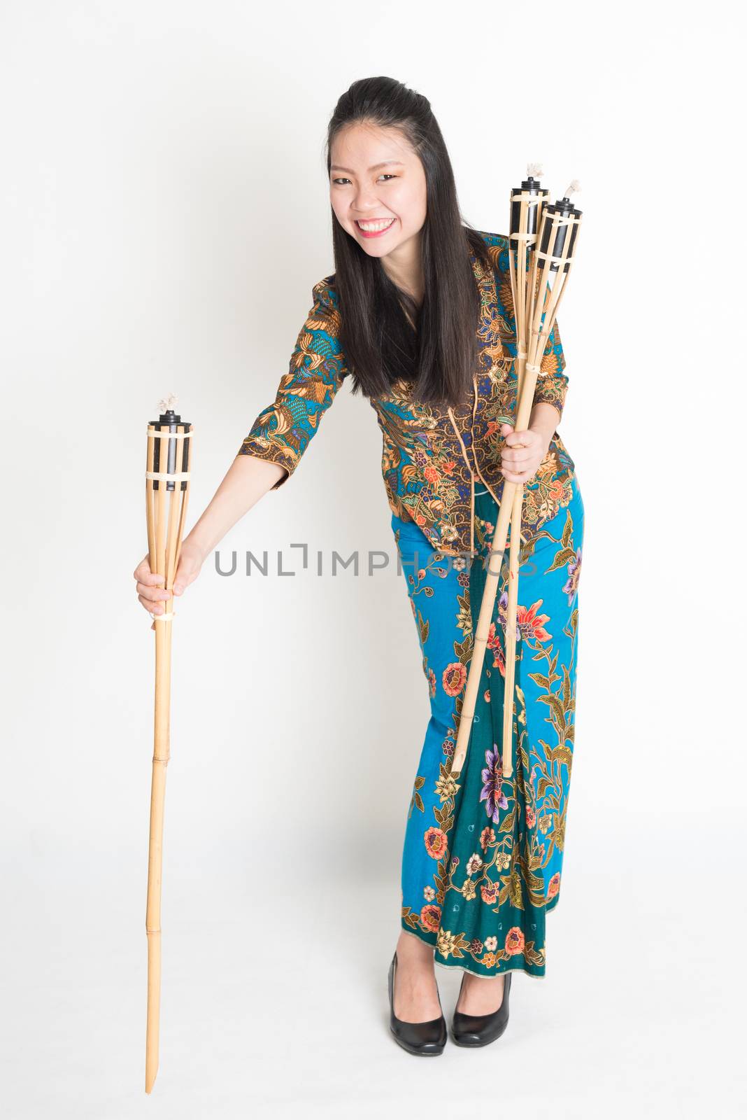 Full body portrait of Southeast Asian woman in batik dress hands holding tiki torch standing on plain background.