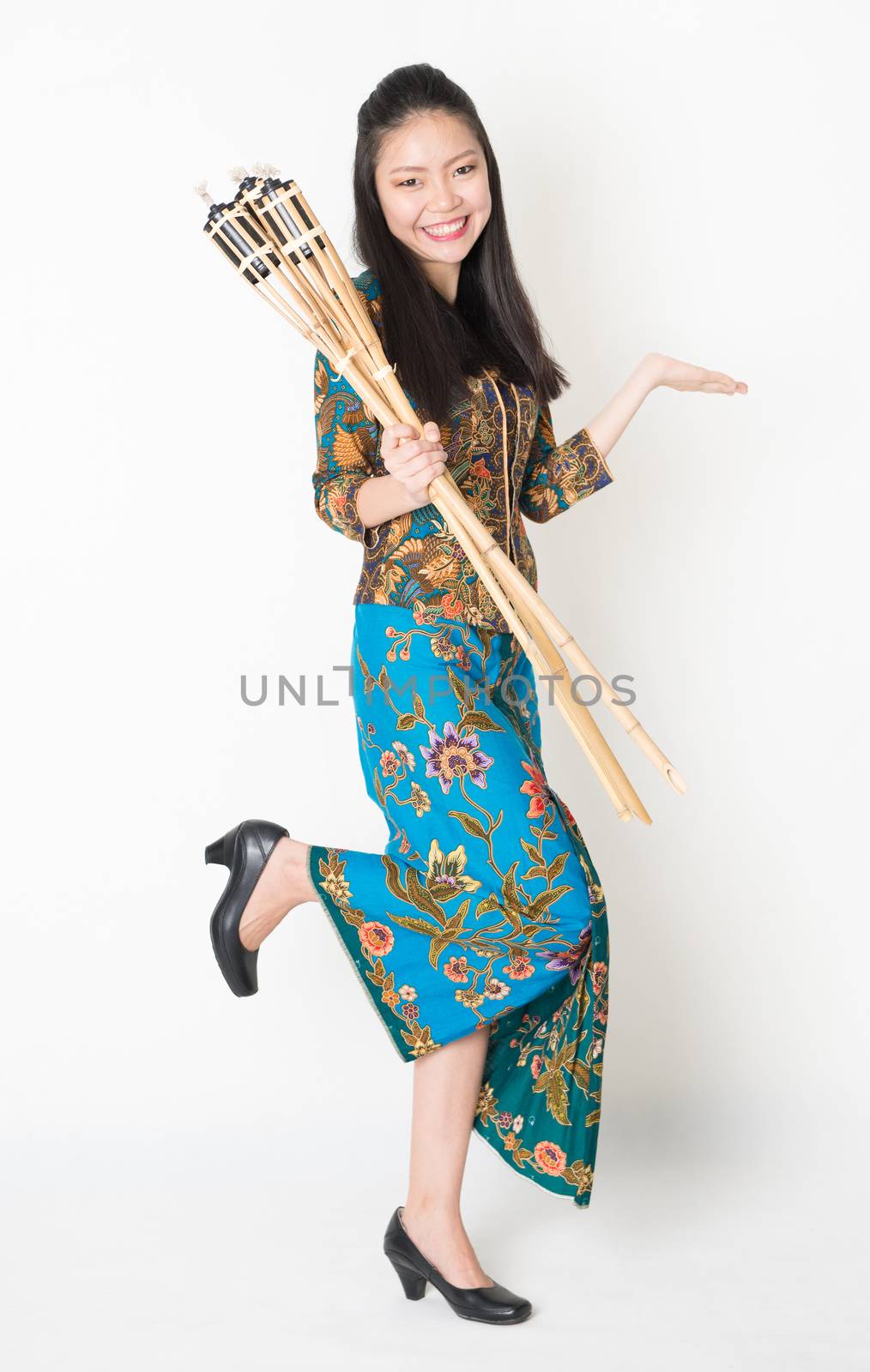 Full body portrait of Southeast Asian woman in batik dress hand holding bamboo oil lamp showing something standing on plain background.