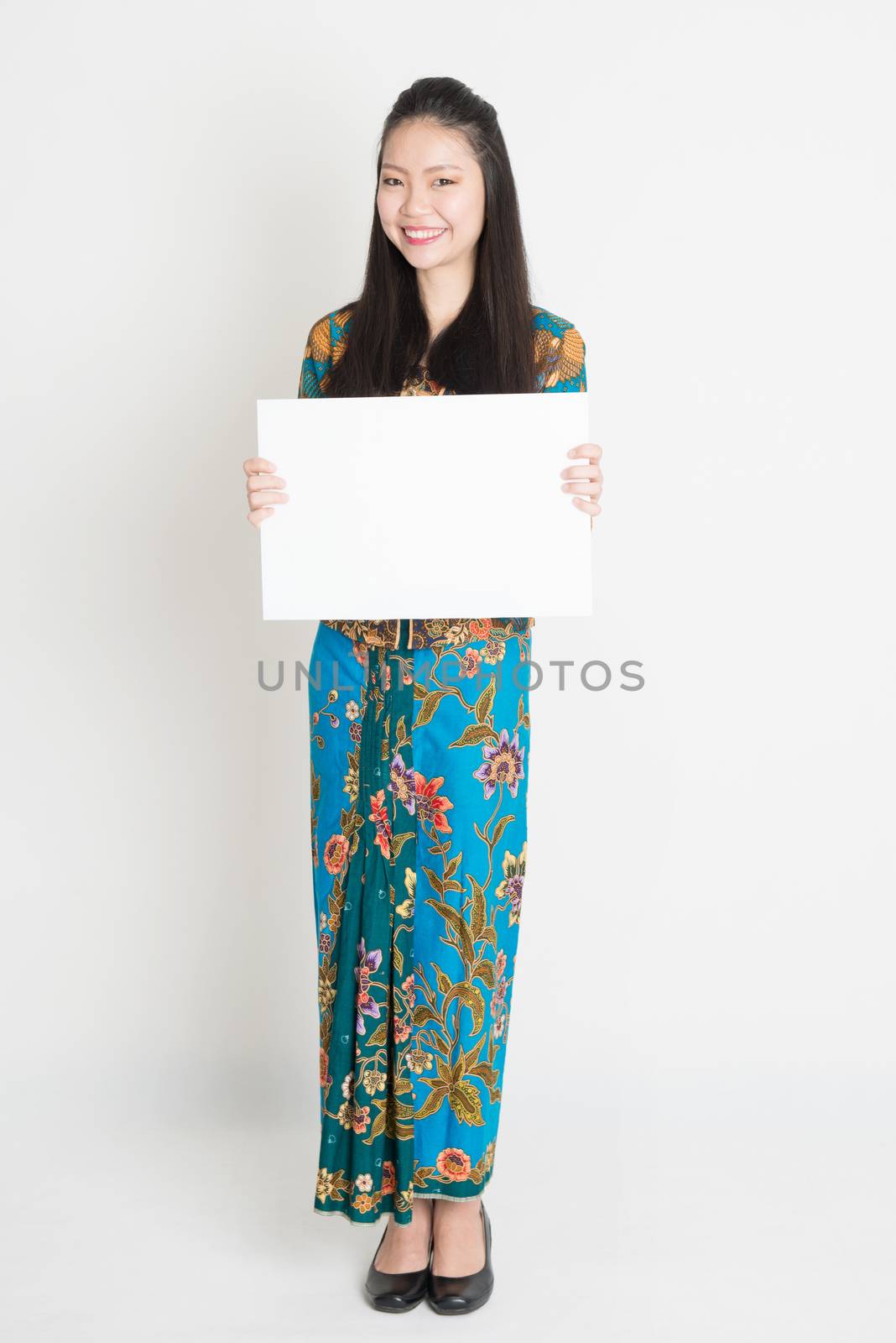 Full body portrait of Southeast Asian girl in batik dress hands holding white blank board, standing on plain background.
