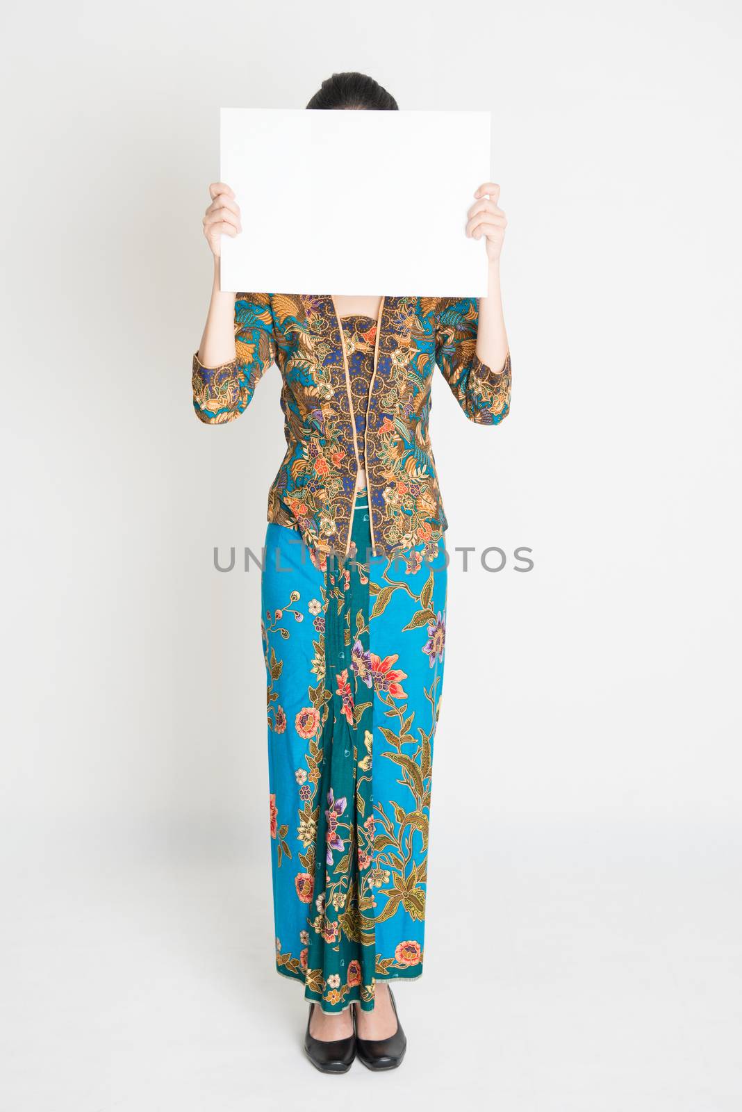 Full length Southeast Asian girl in batik dress holding a white card covering face standing on plain background.