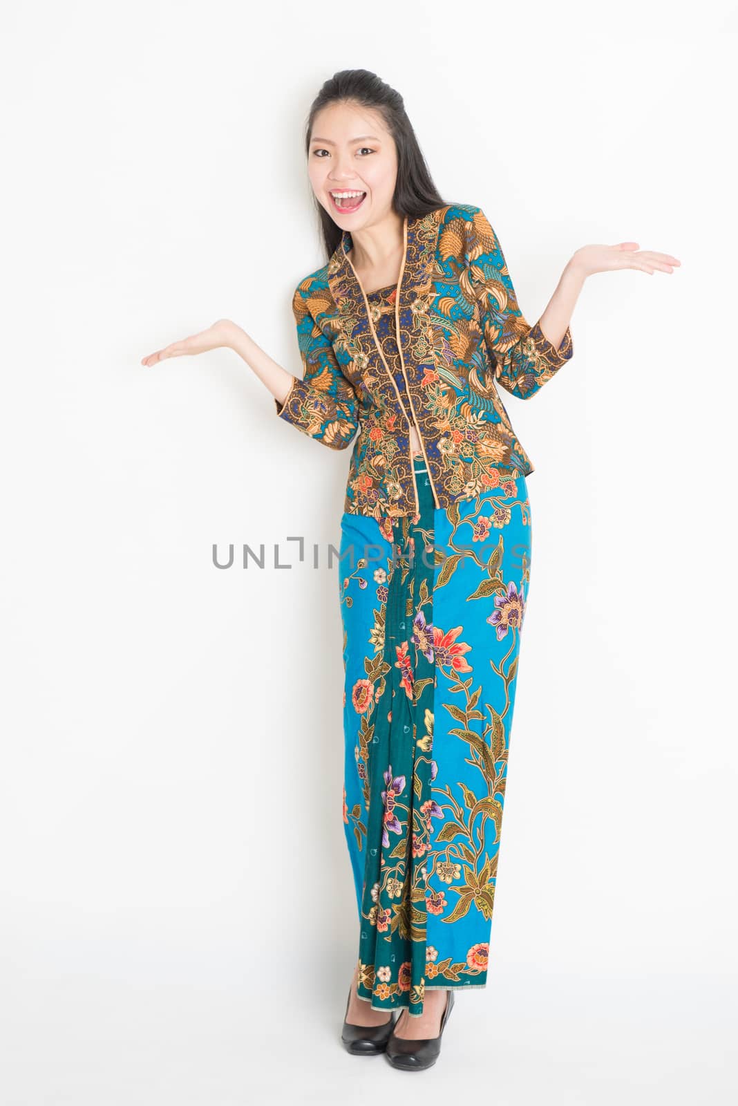 Full body portrait of cheerful Southeast Asian woman in batik dress hands showing something, standing on plain background.