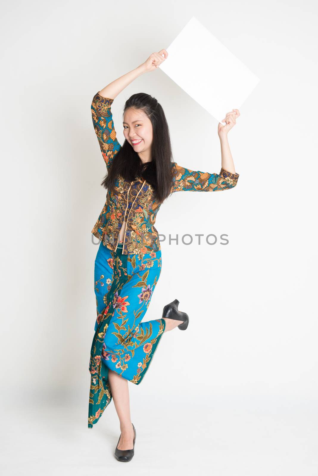 Full body portrait of Southeast Asian girl in batik dress hands holding white blank card jumping around on plain background.