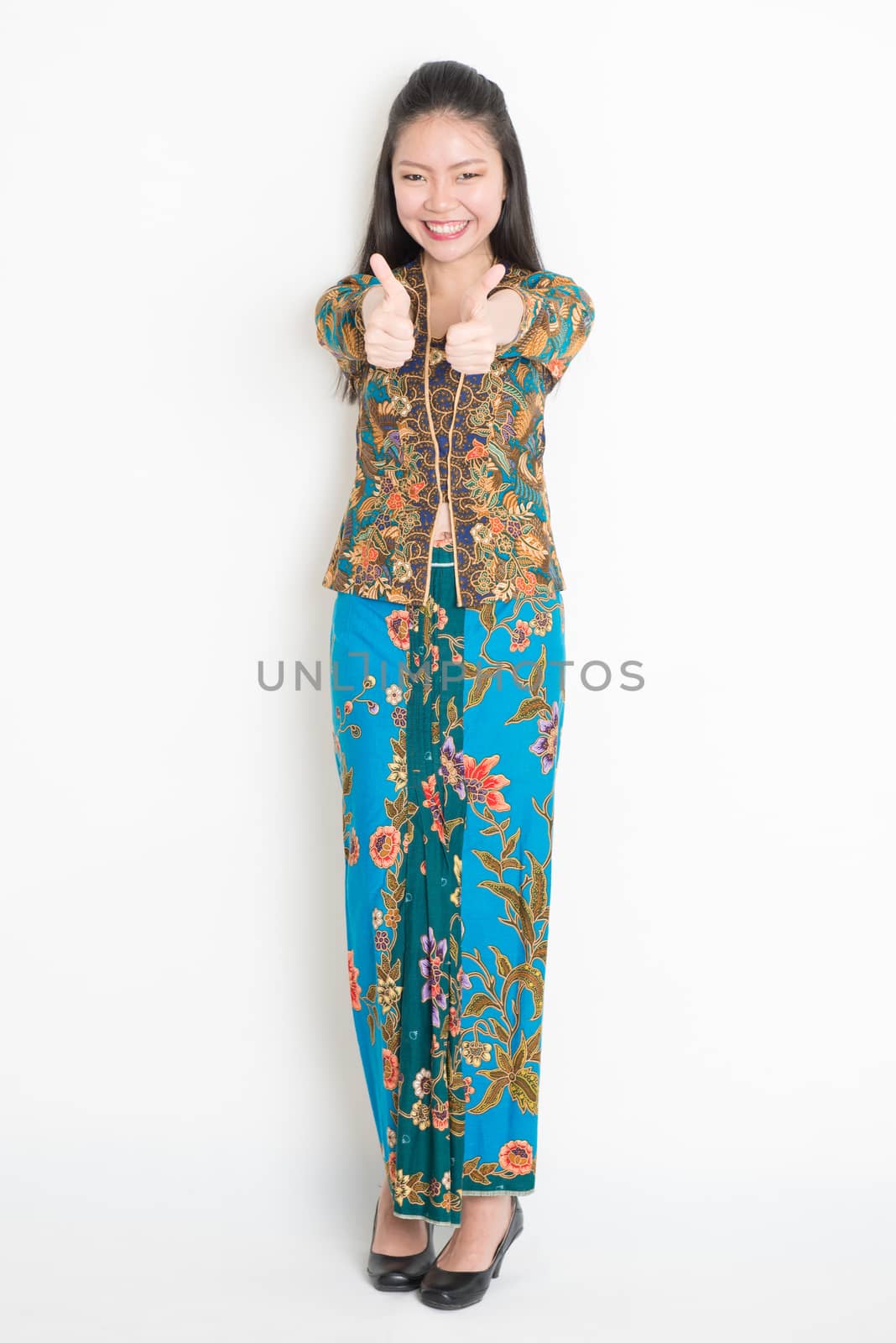 Full length portrait of Southeast Asian woman in batik dress thumbs up standing on plain background.