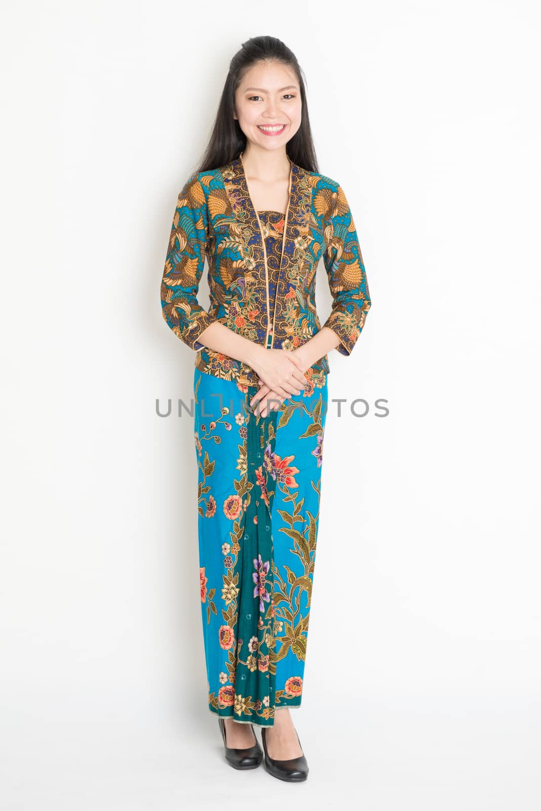 Full length Southeast Asian girl in batik dress standing on plain background.