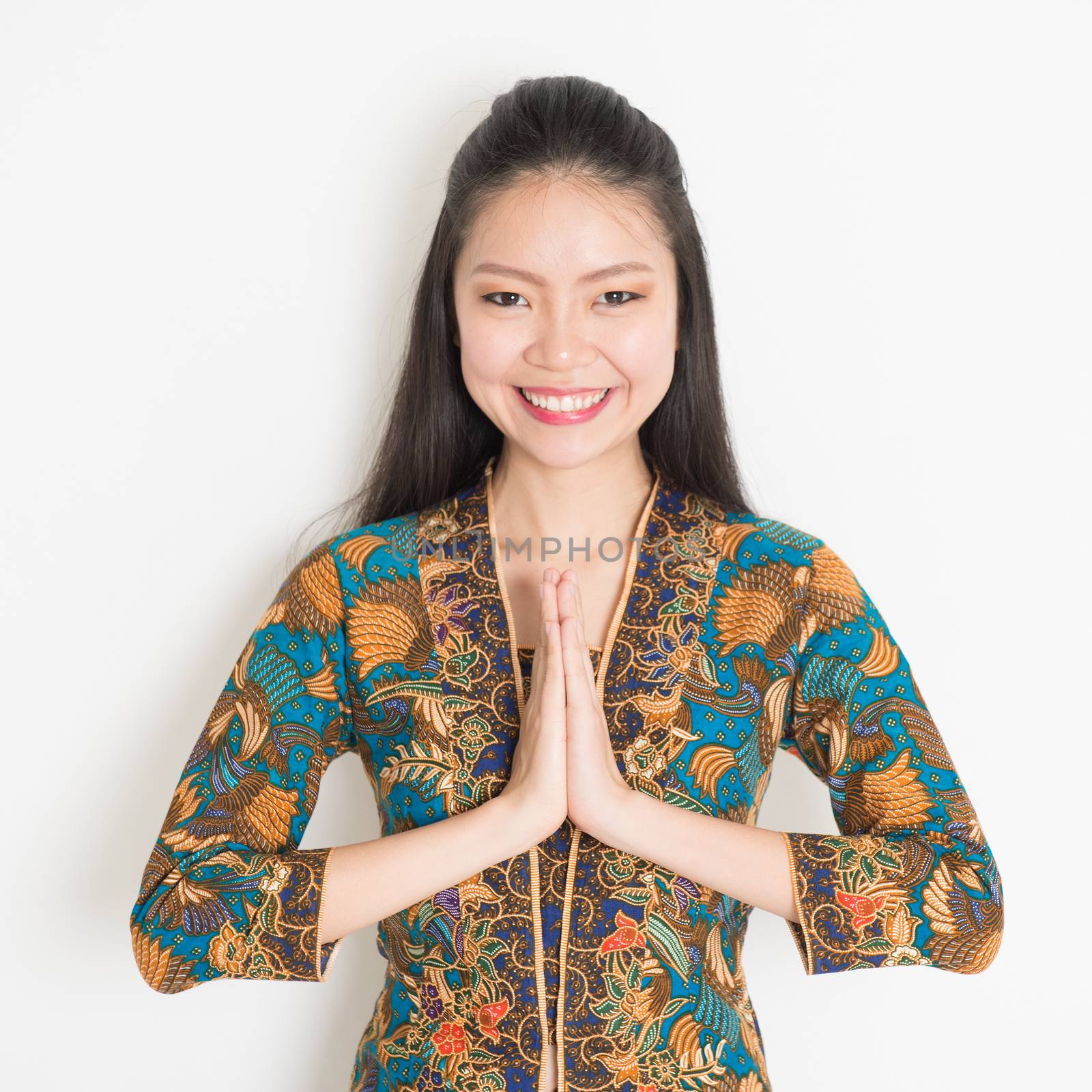 Portrait of happy Southeast Asian woman with batik dress in greeting gesture on plain background.