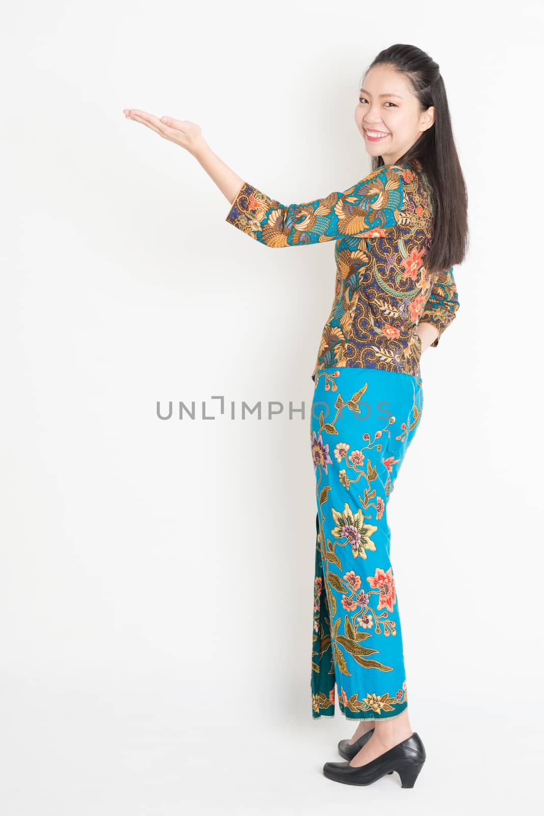 Full length portrait of Southeast Asian female in batik dress hand holding something standing on plain background.