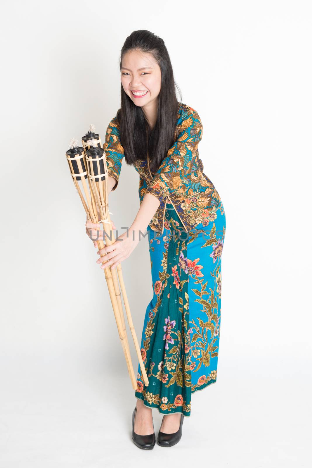 Full body portrait of Southeast Asian woman in batik dress hands holding pelita standing on plain background.