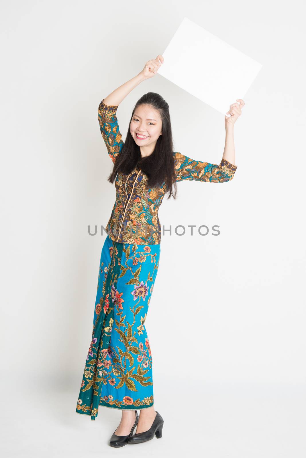 Full body portrait of Southeast Asian girl in batik dress hands holding white blank poster standing on plain background.