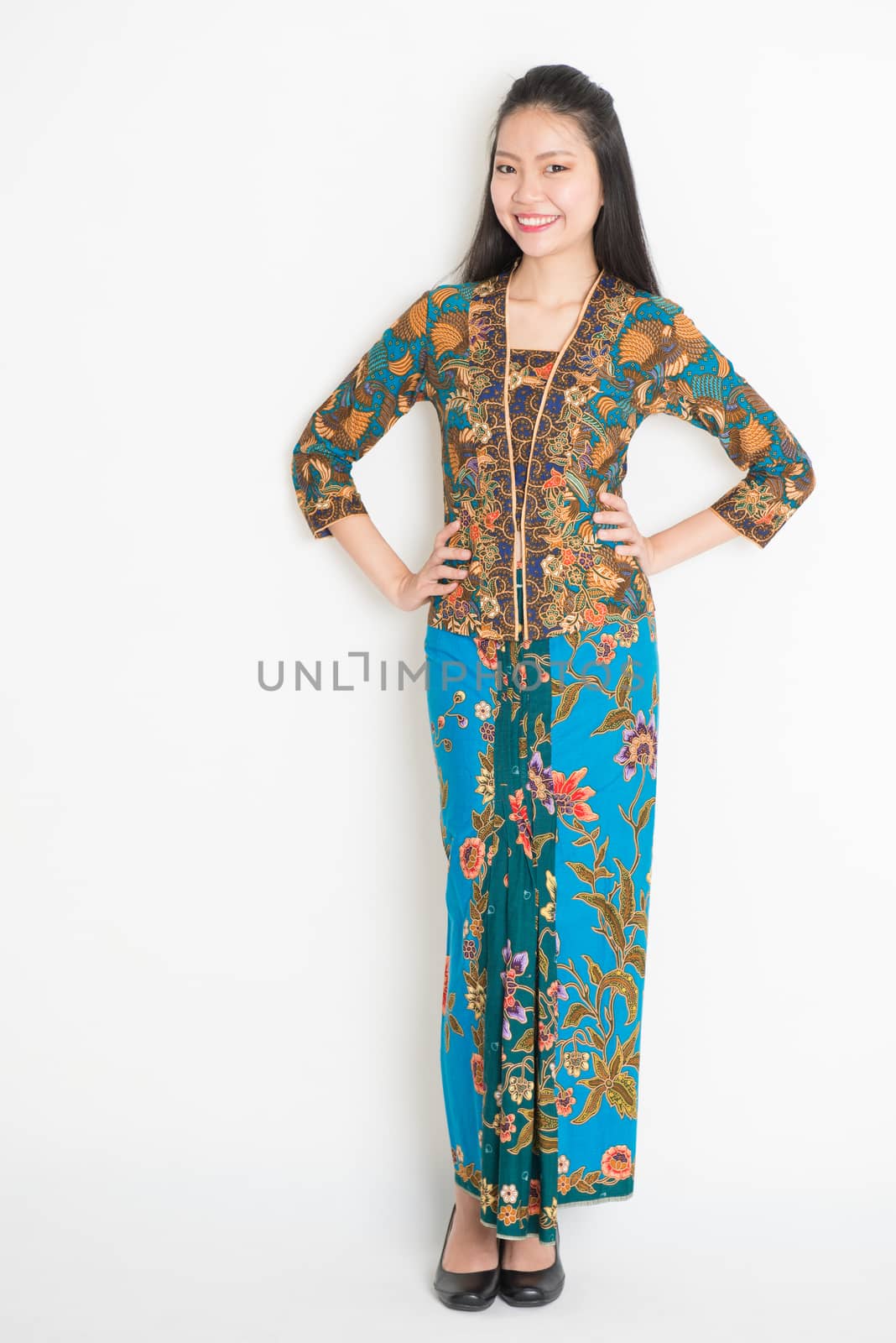 Full body Southeast Asian woman in batik dress standing on plain background.