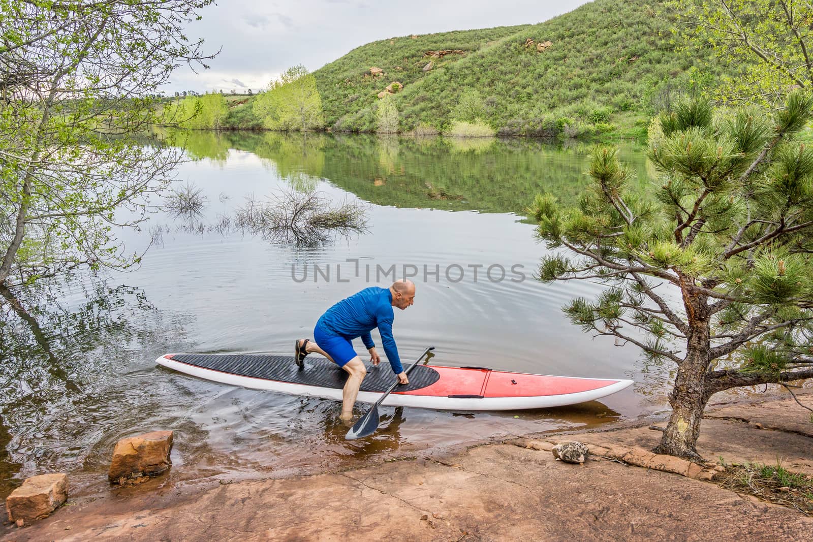 senior male paddler launching his red SUP paddleboard at a rocky lake shore (Horsetooth Reservoir, Fort Collins, Colorado)