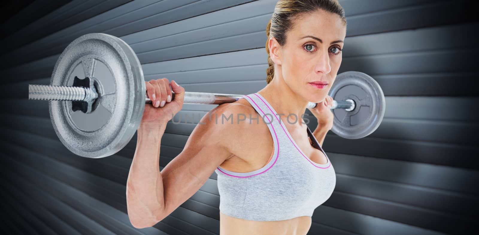 Strong female crossfitter lifting barbell behind head looking at camera against grey shutters