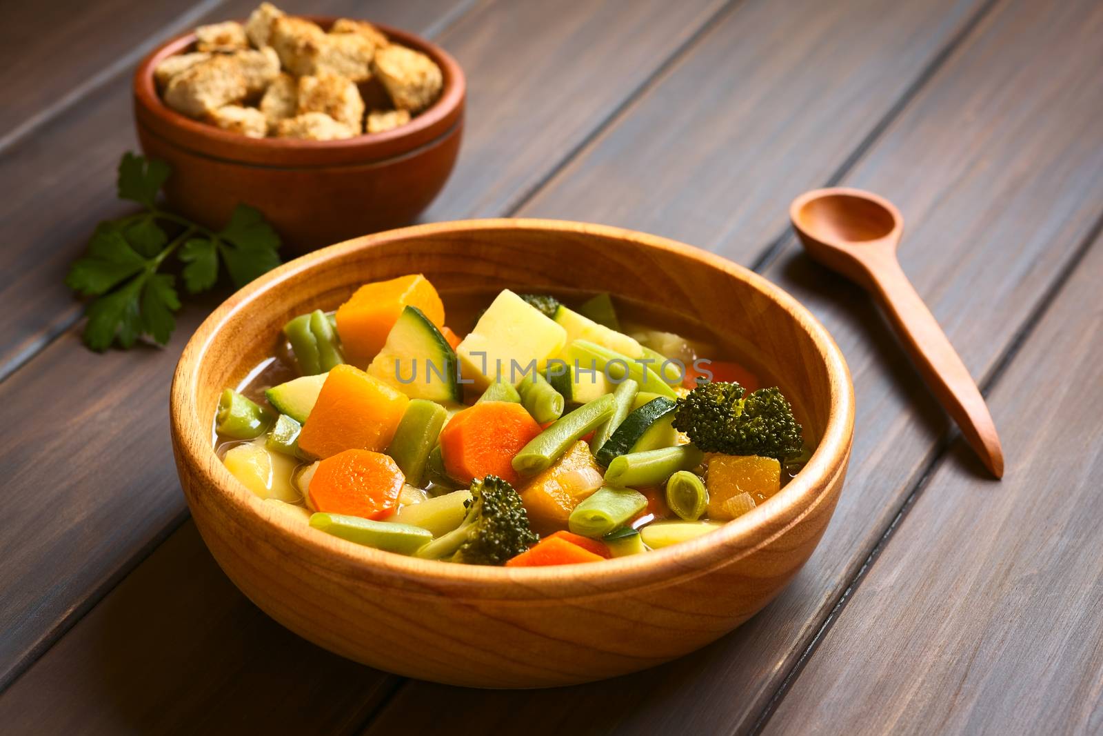 Wooden bowl of vegetable soup made of zucchini, green bean, carrot, broccoli, potato and pumpkin with a small bowl of croutons in the back, photographed with natural light (Selective Focus, Focus one third into the soup)