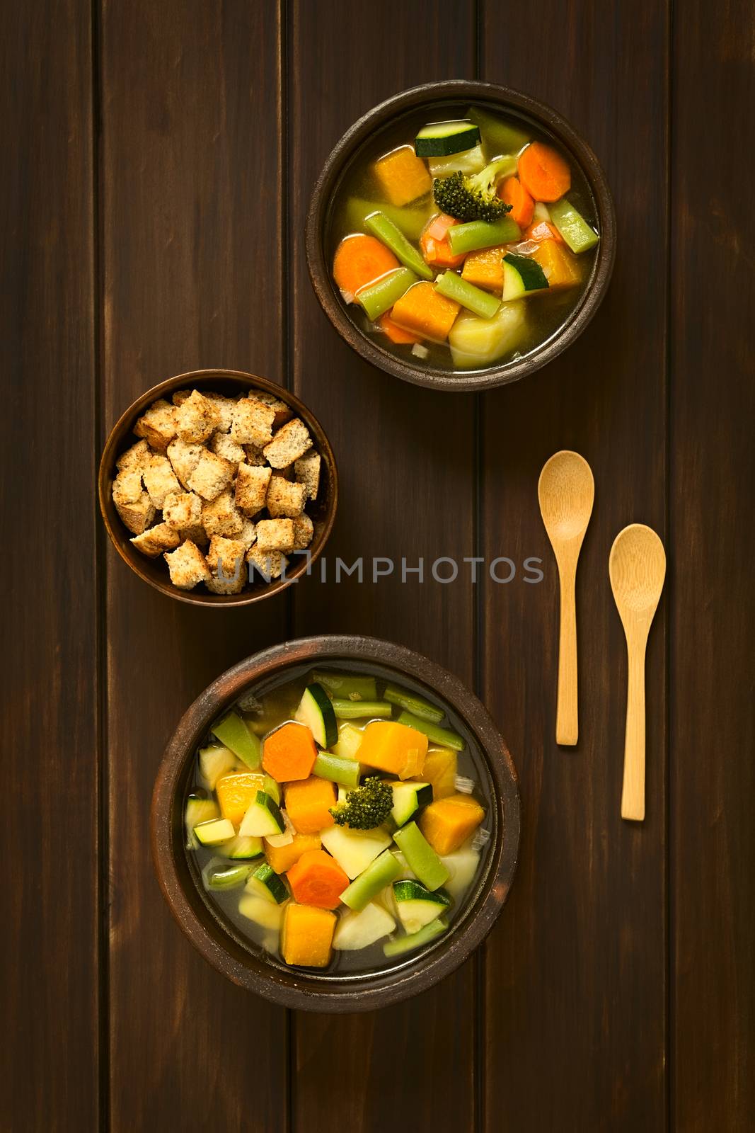 Overhead shot of two rustic bowls of vegetable soup made of zucchini, green bean, carrot, broccoli, potato and pumpkin with a small bowl of croutons, photographed on dark wood with natural light