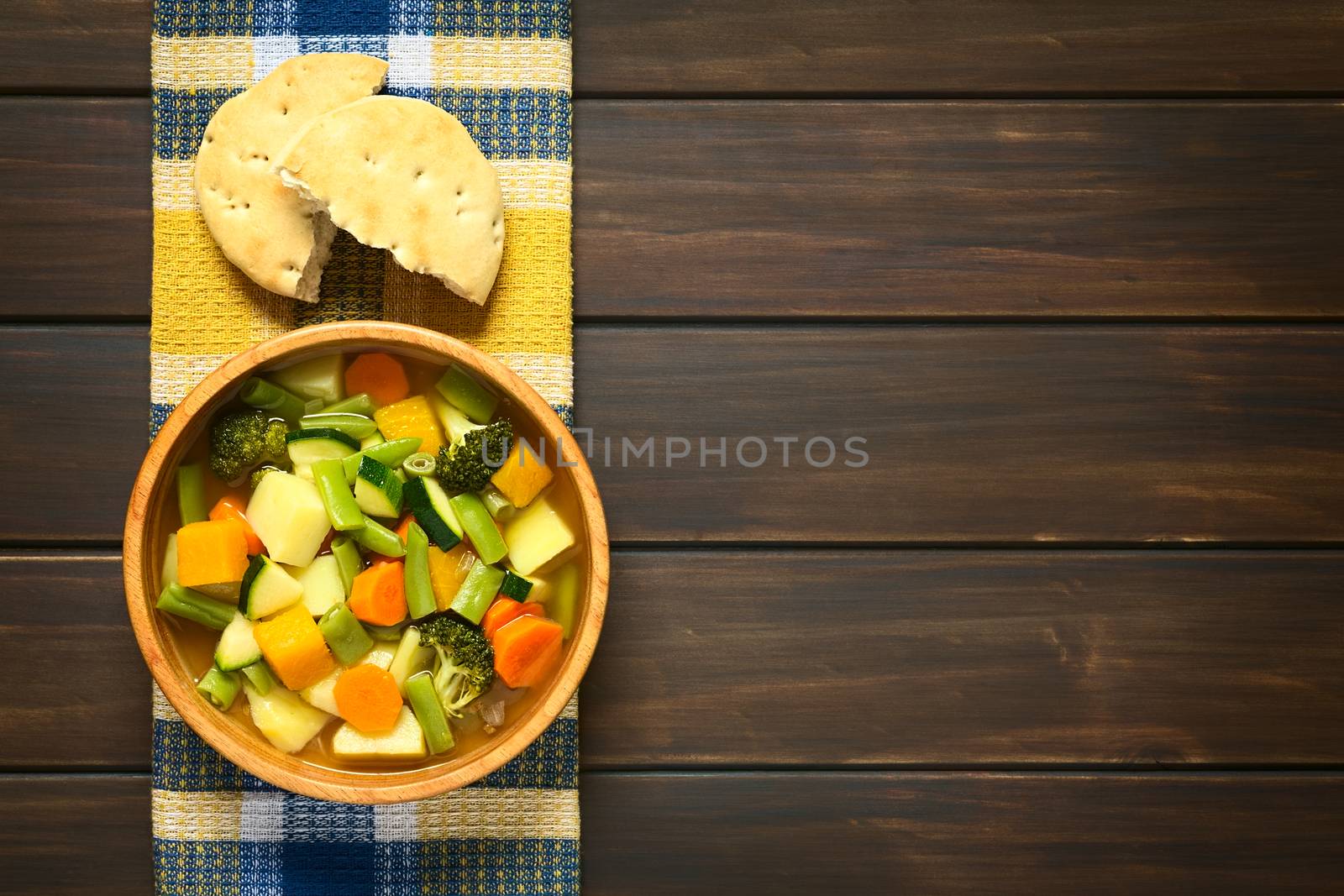 Wooden bowl of vegetable soup made of zucchini, green bean, carrot, broccoli, potato and pumpkin with a bun on kitchen towel, photographed on dark wood with natural light