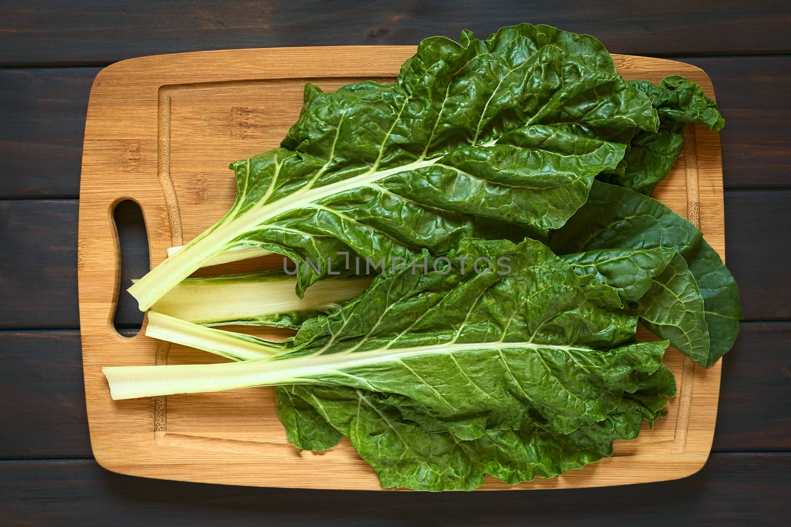 Overhead shot of raw chard leaves (lat. Beta vulgaris) on wooden board, photographed on dark wood with natural light