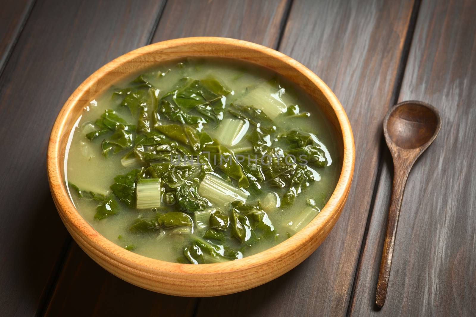 Chard soup in wooden bowl with a small wooden spoon, photographed on dark wood with natural light (Selective Focus, Focus one third into the soup)