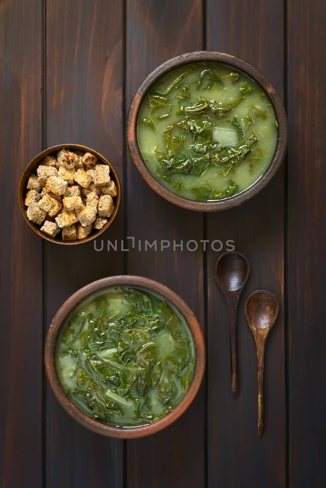 Overhead shot of two rustic bowls of chard soup and a small bowl of croutons with two wooden spoons, photographed on dark wood with natural light