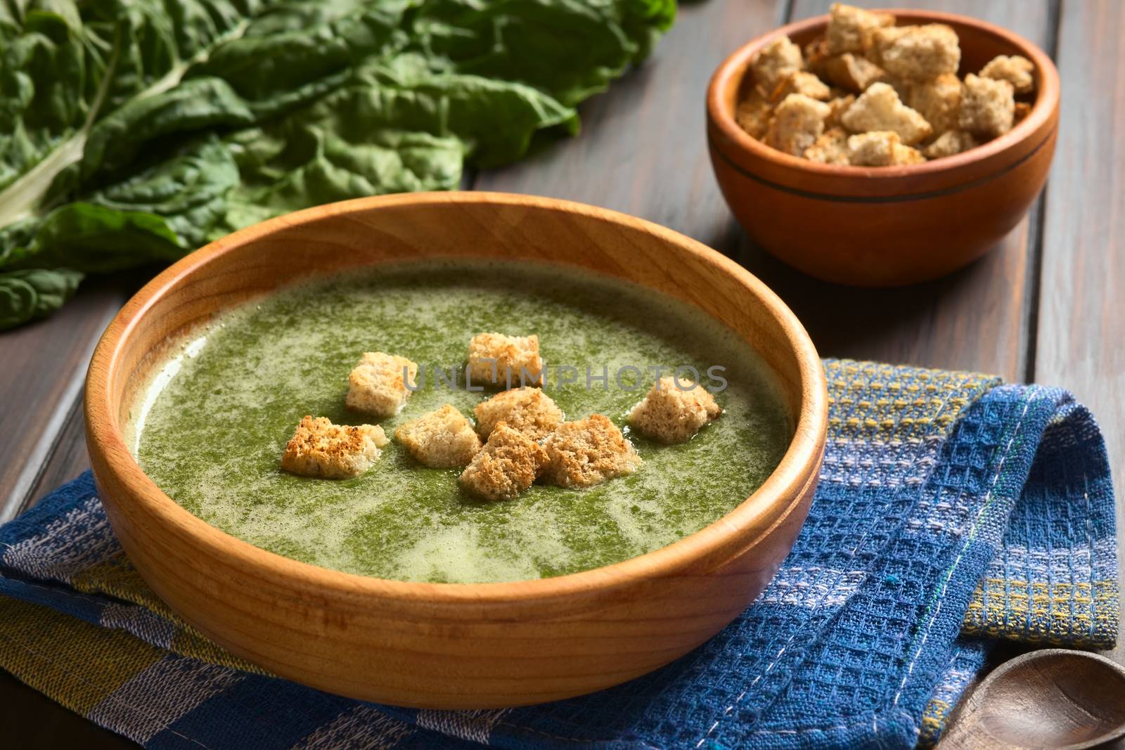 Cream of chard soup with croutons in wooden bowl, fresh raw chard leaves and a small bowl of croutons in the back, photographed on dark wood with natural light (Selective Focus, Focus on the first croutons)