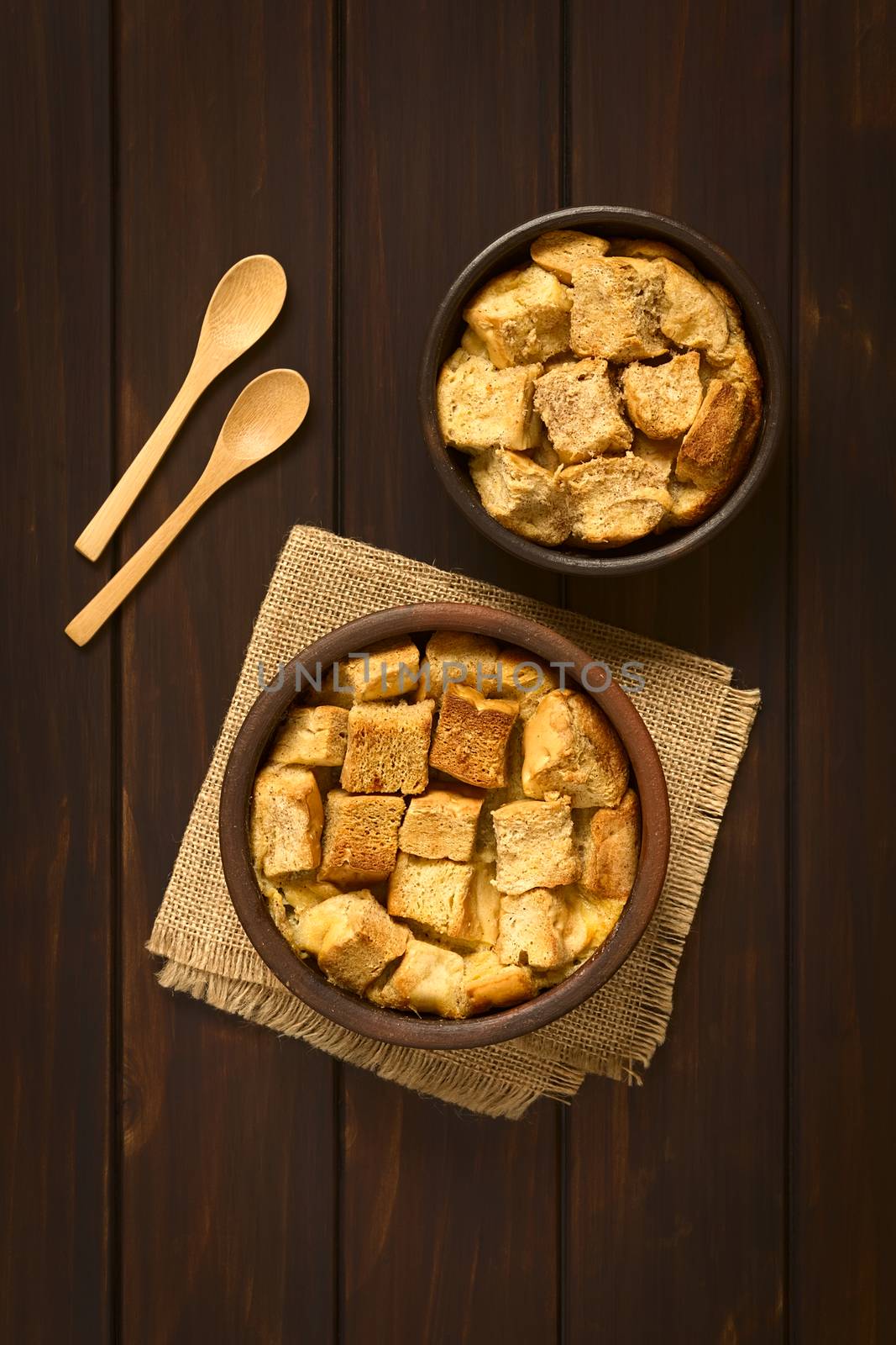 Overhead shot of two rustic bowls of bread pudding made of diced stale bread, milk, egg, cinnamon, sugar and butter, photographed on dark wood with natural light