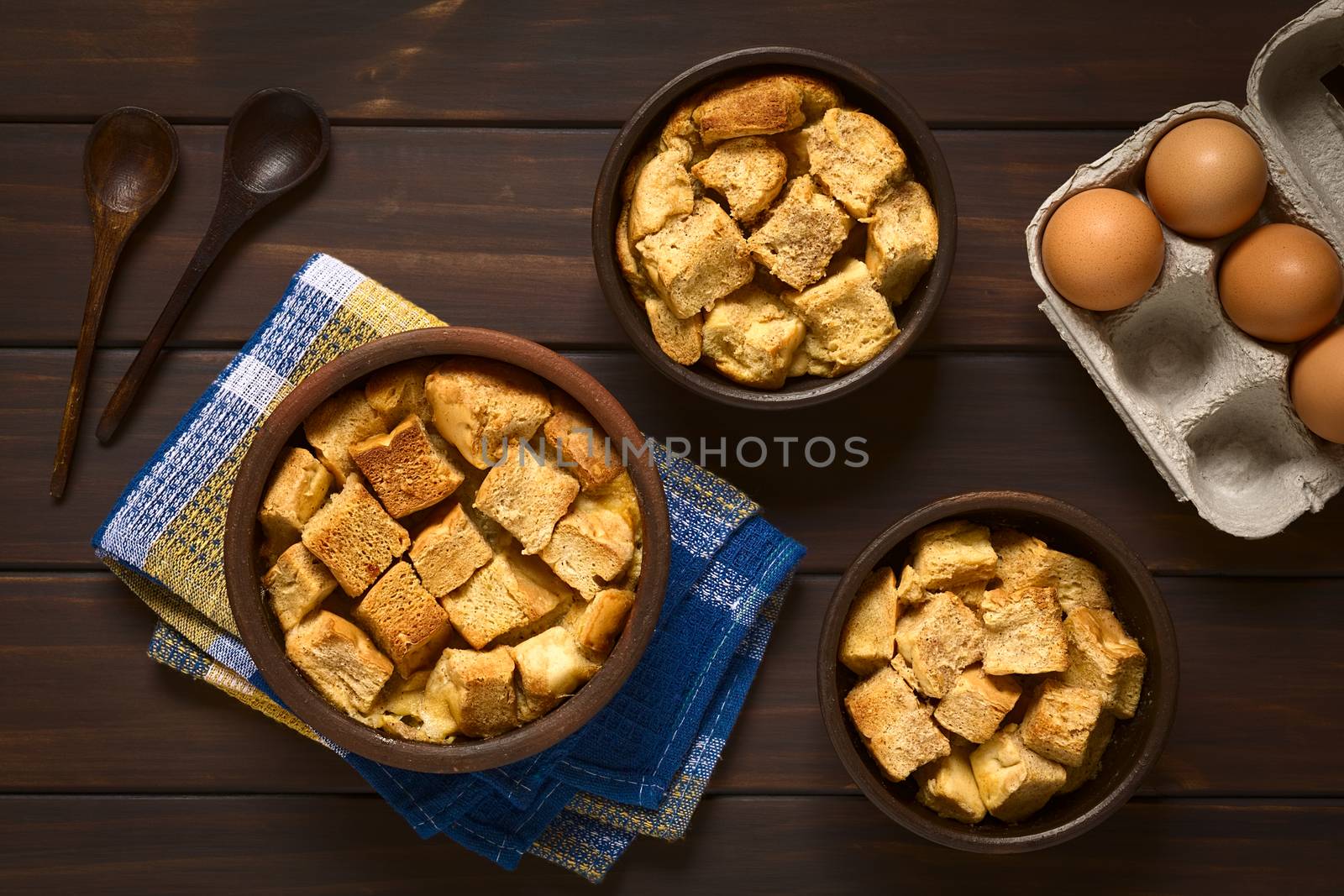 Overhead shot of three rustic bowls of bread pudding made of diced stale bread, milk, egg, cinnamon, sugar and butter, photographed on dark wood with natural light