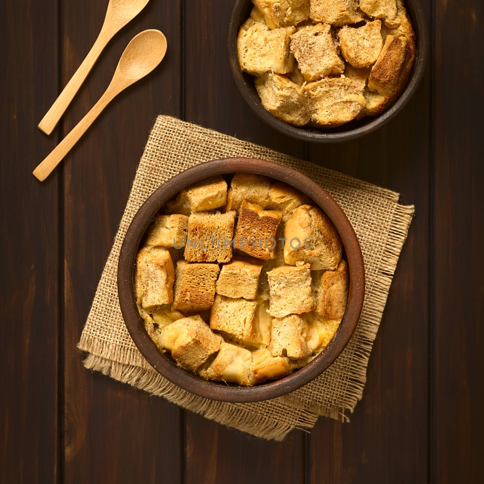 Overhead shot of two rustic bowls of bread pudding made of diced stale bread, milk, egg, cinnamon, sugar and butter, photographed on dark wood with natural light