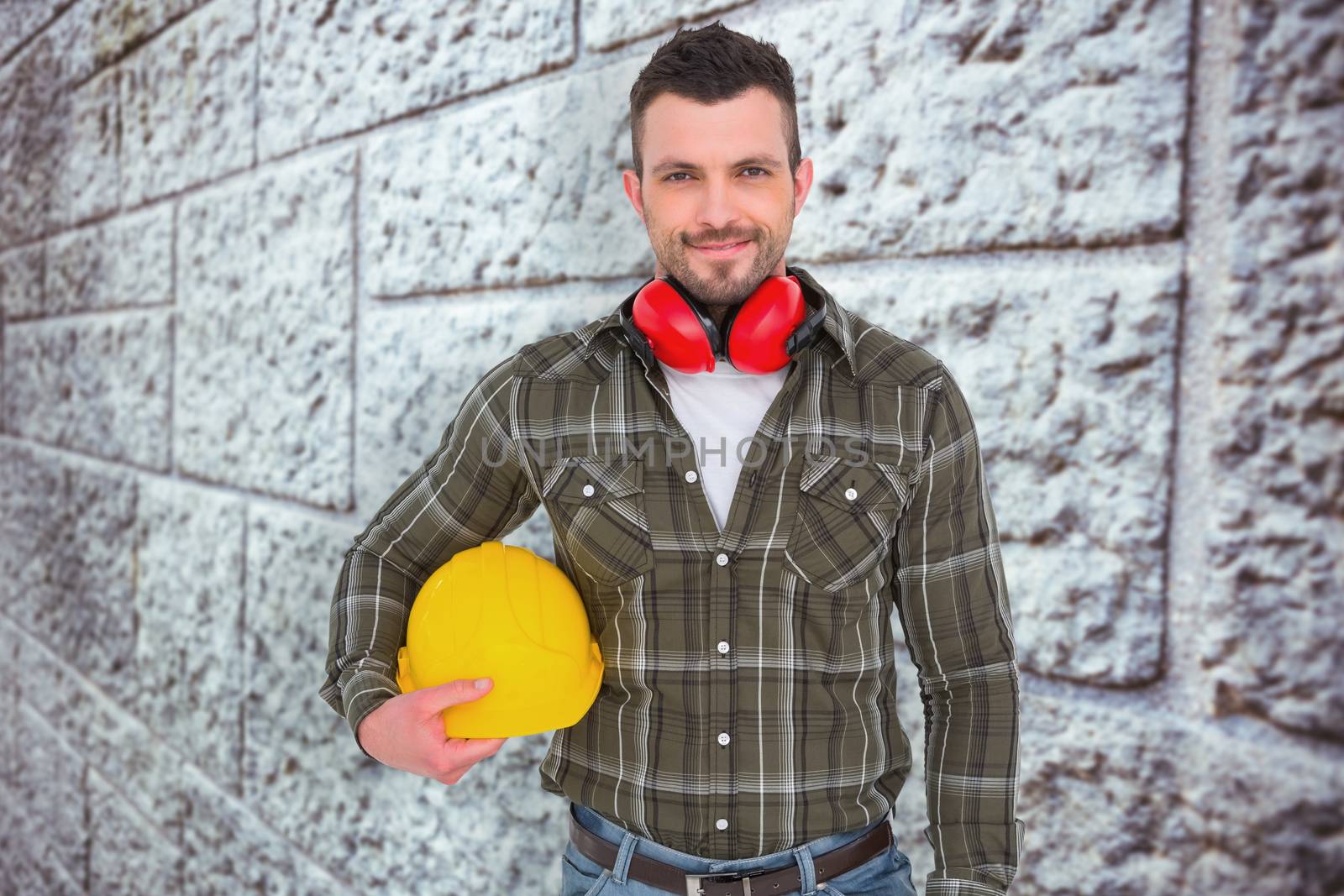 Handyman with earmuffs holding helmet  against grey brick wall