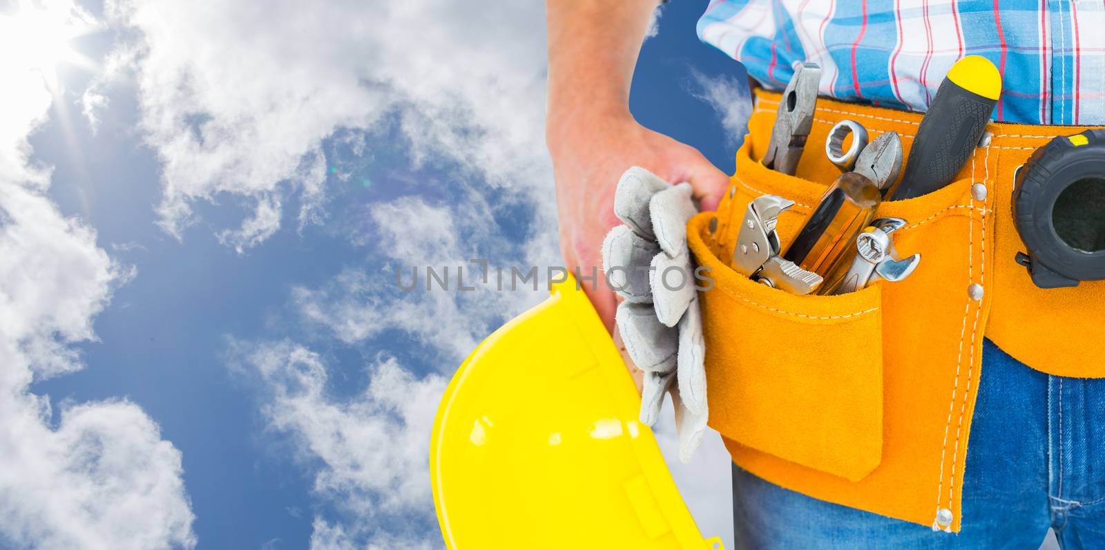 Manual worker wearing tool belt while holding gloves and helmet against bright blue sky with clouds