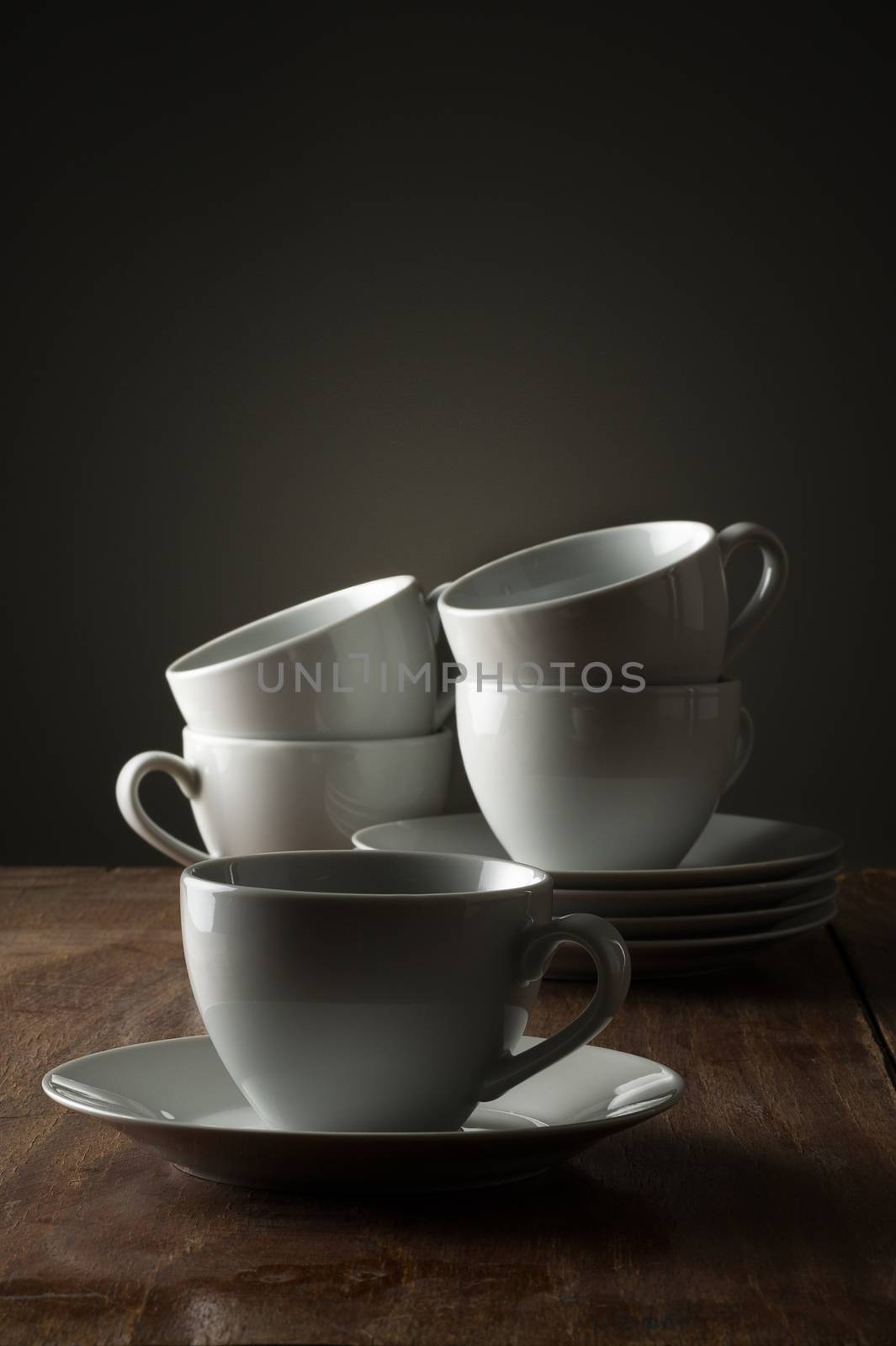 Five plain white ceramic coffee or tea cups on a wooden kitchen counter or table side lit with dramatic lighting with a single cup and saucer in the foreground