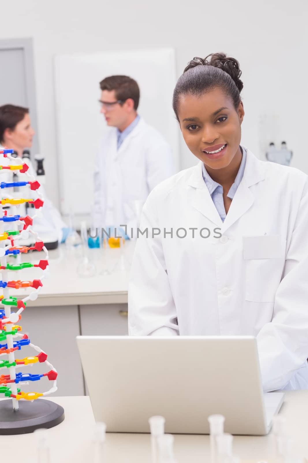 Smiling scientist using laptop while colleagues talking together  by Wavebreakmedia