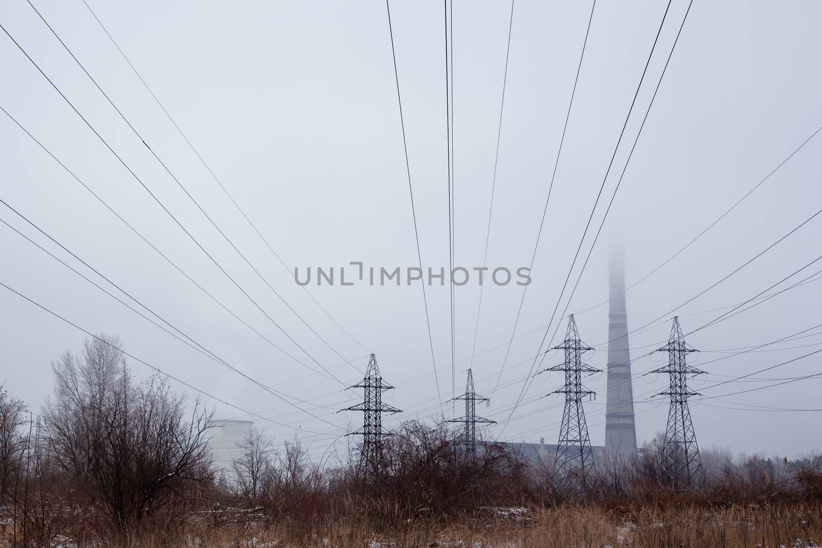 Electricity pylons, power lines, smoke stack and cooling tower of the industrial plant in the foggy winter day.
