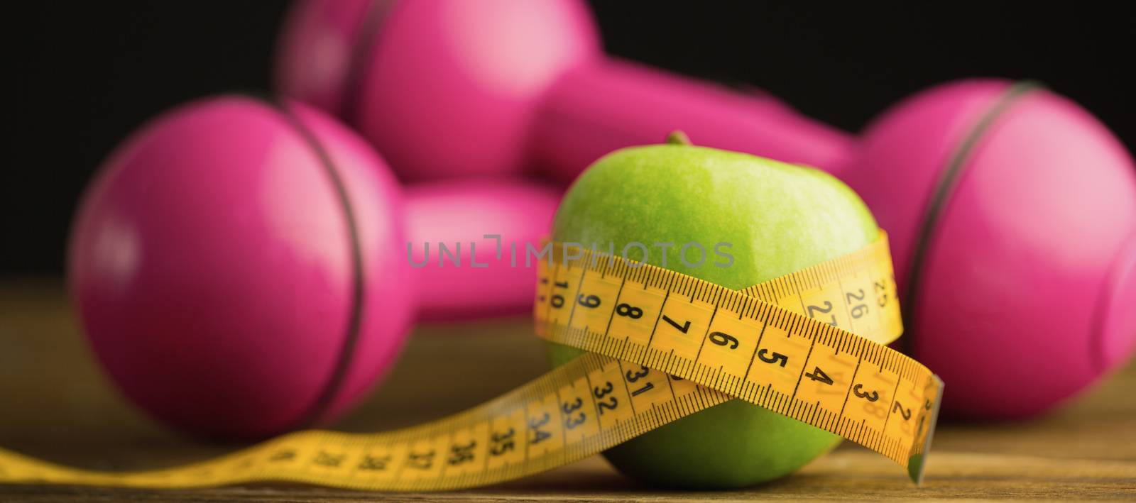 Pink dumbbells with green apple and measuring tape on wooden background