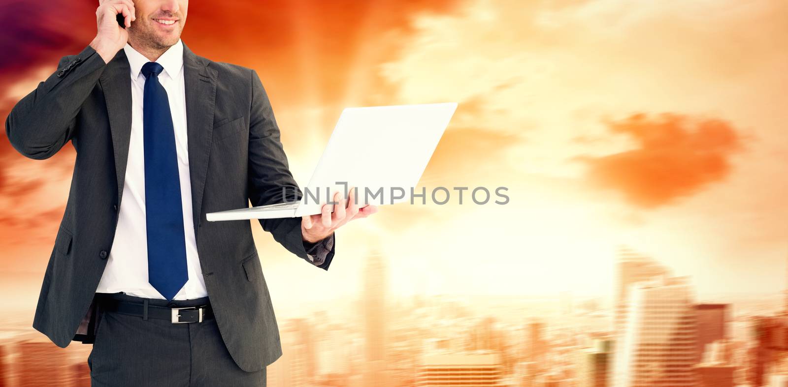 Businessman talking on phone holding laptop against sun shining over city