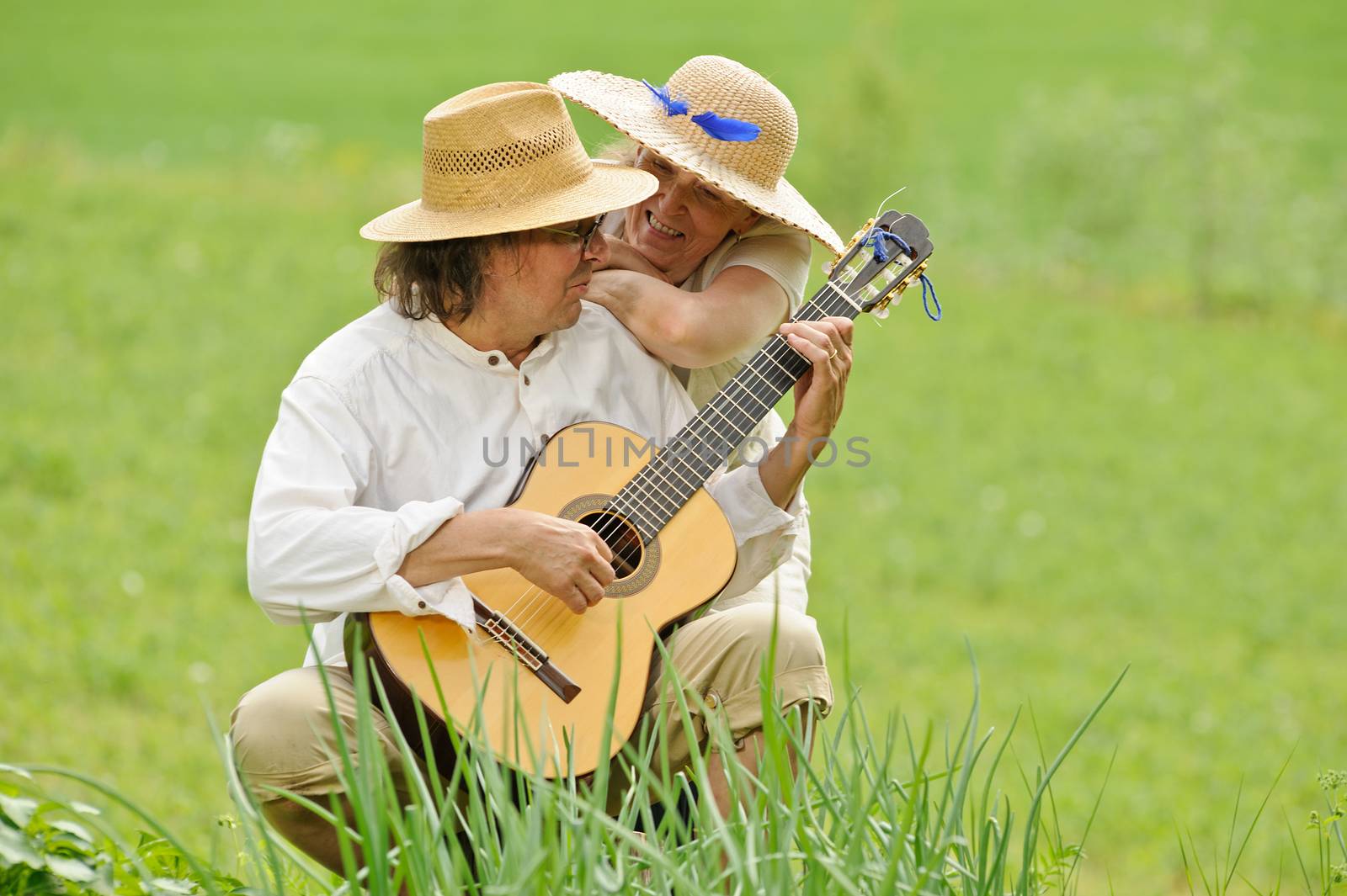Senior couple having fun together. The man is sitting and playing a guitar. The woman hugs him from behind. They are outdoors.