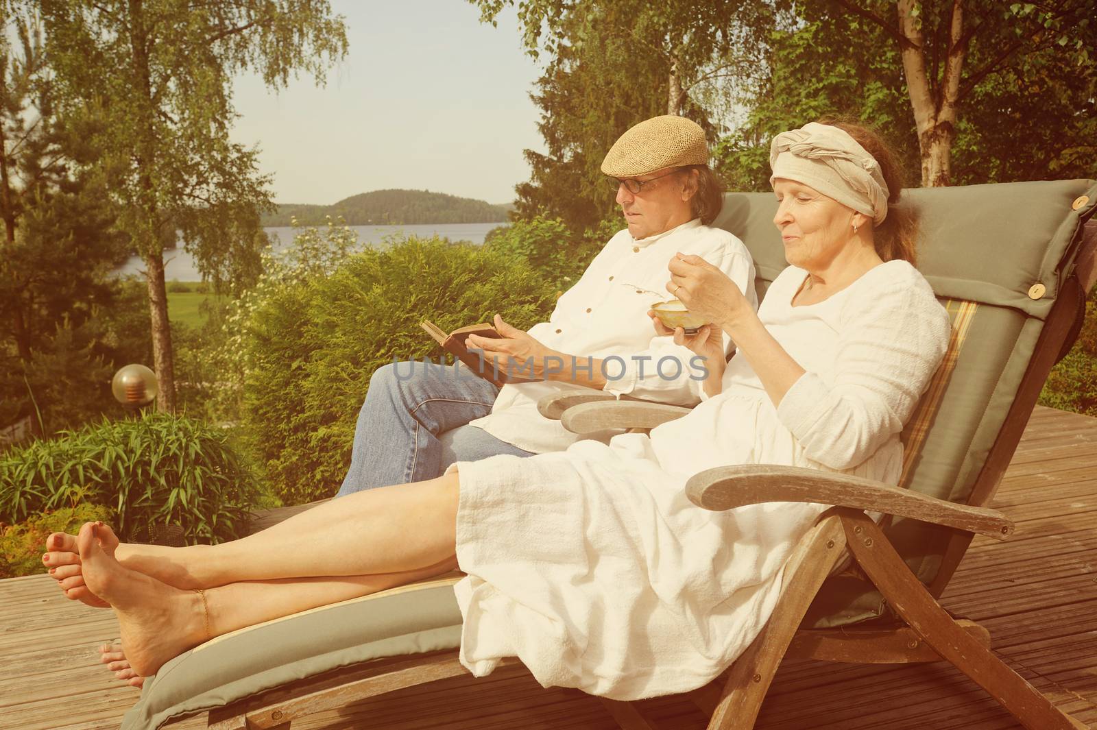 Senior couple relax in deckchairs on a wooden terrace, She is eating from a bowl and he is reading a book. Digital filters used to create a retro look. Lake and forest in the background.