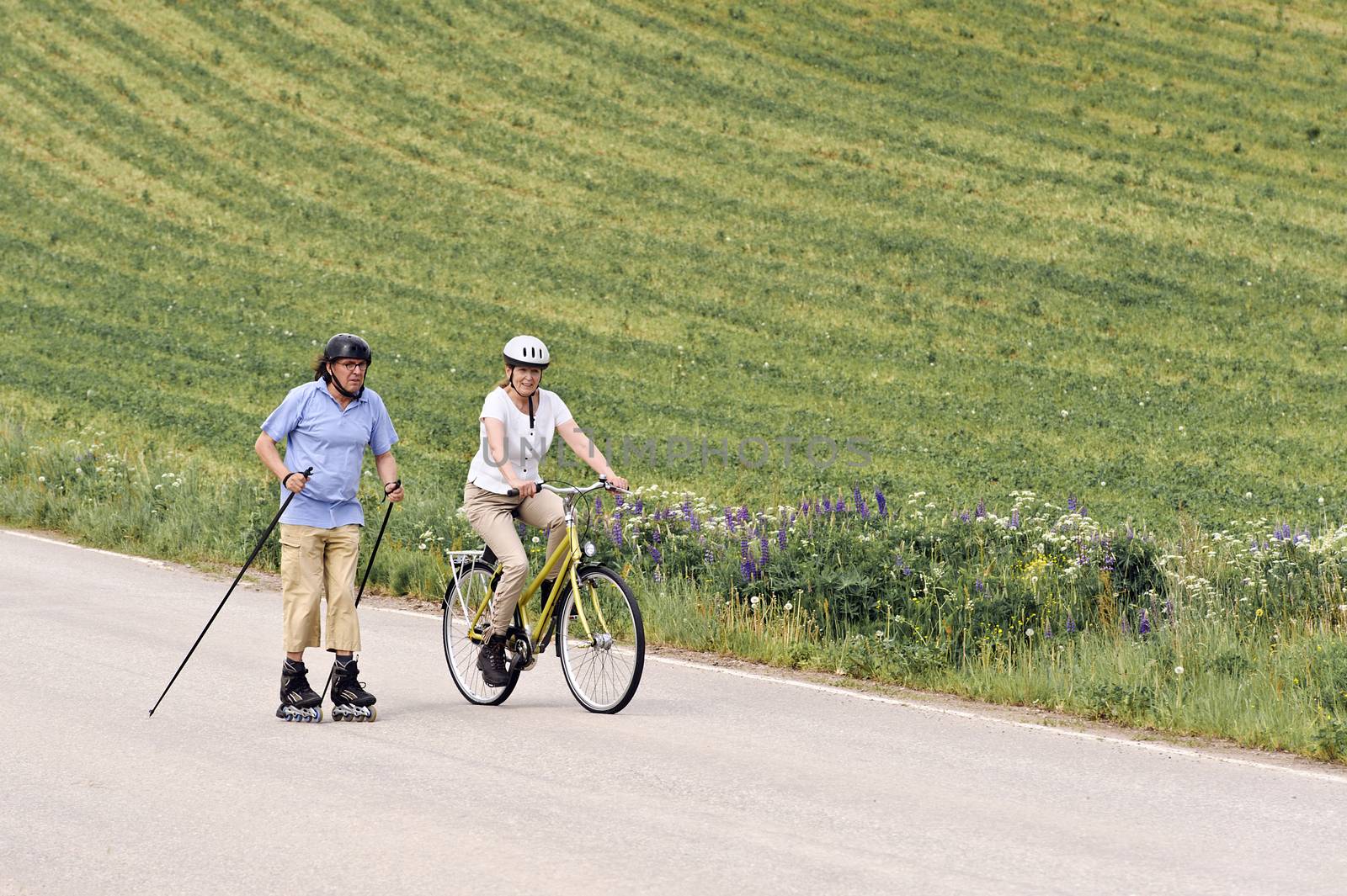 Senior couple vigorously exercising. The man is Nordic inline skating, and the woman is cycling. They're on a country road through green fields.