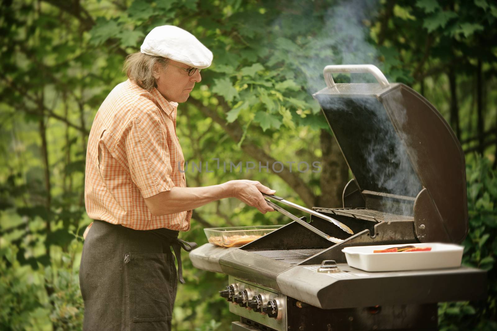 Senior man grilling outdoors. Smoke rises from the barbeque. The picture is desaturated and has been treated with digital filters