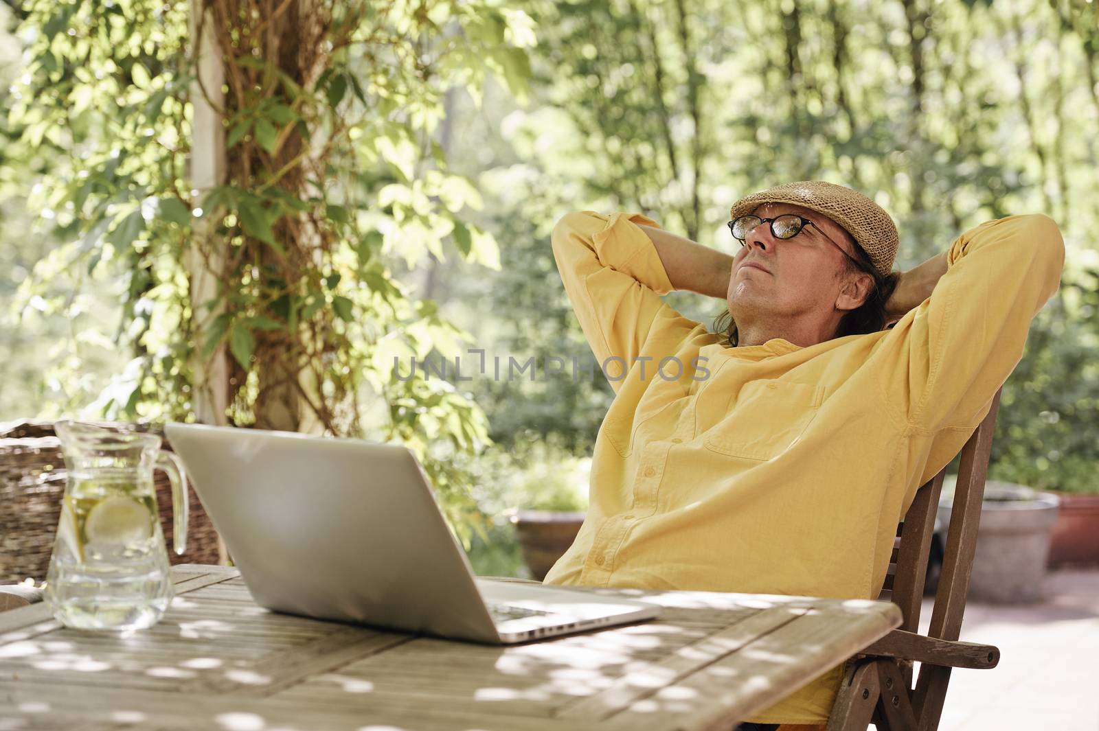 Senior man sits outdoors at a wooden table under a pergola and works on a laptop computer. It's summer and there's a background of green trees and bushes