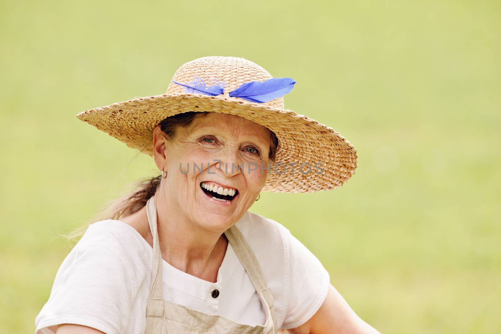 Senior woman wearing a straw hat  is looking at teh camera and laughing. The bacground is a blurred green field