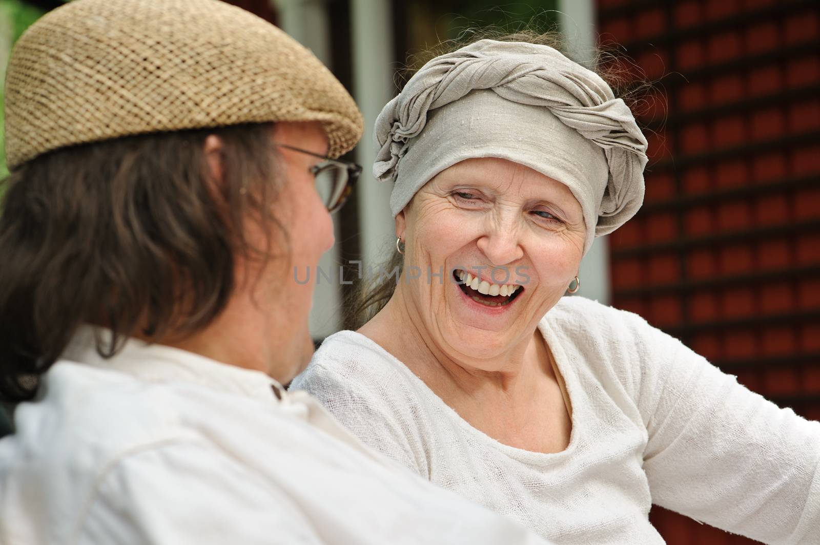 Senior man and woman sitting outdoors. The woman laughs at something the man has said. Window and red wall in the background