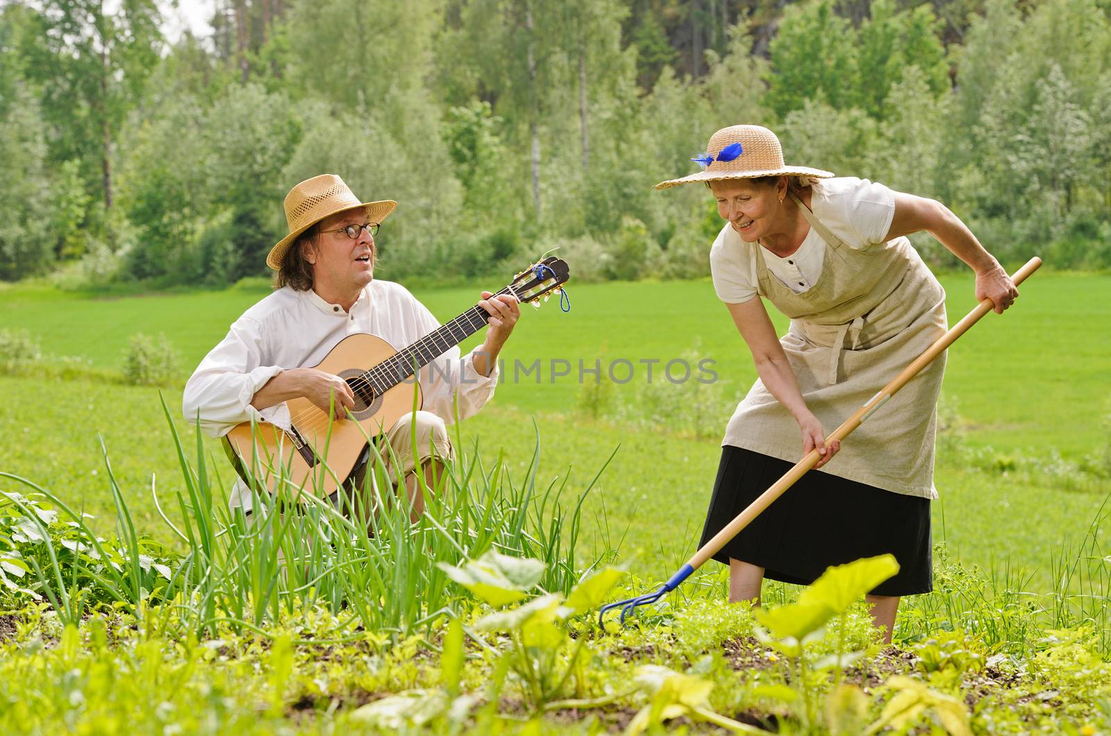 Senior man and woman in a vegetable garden. The woman is raking the soil. The man is playing a guitar