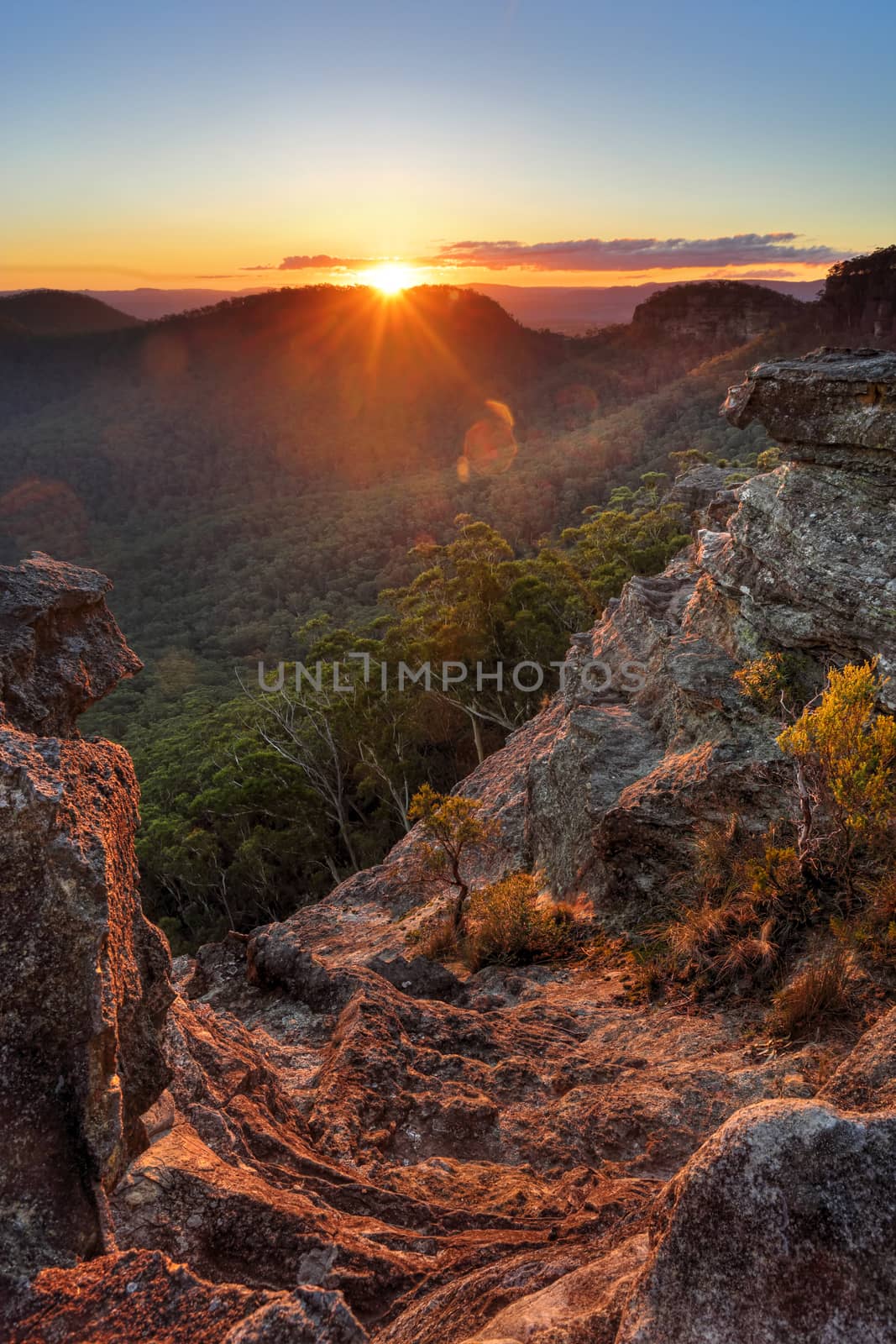 The last rays of sunshine stream over Wilsons Gully and the valley and highlight the foreground cliffs and rock ledges and foliage.  Sunset views from Sunset Rock, Mt Victoria in the Blue Mountains, Australia