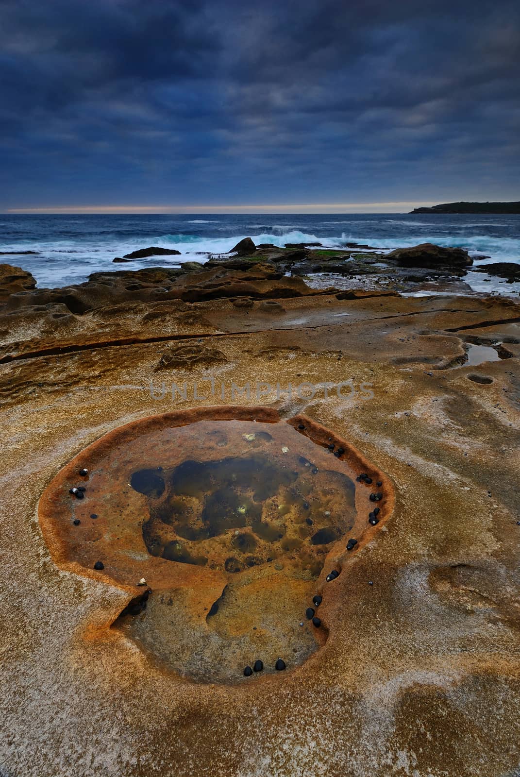 Sunrise seascape with orange rocks and ocean pools and sea shells with cloudy stormy sky and distant cliffs