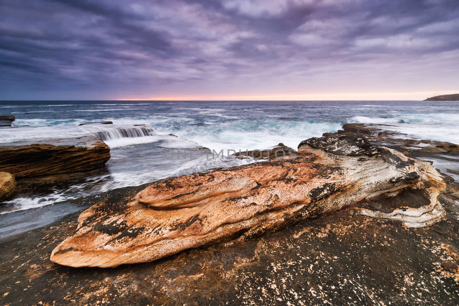 Sunrise seascape with orange rocks and ocean pools and sea shells with cloudy stormy sky and distant cliffs